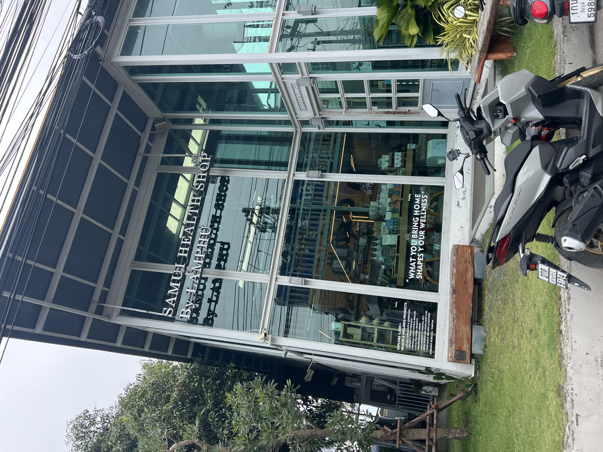 The image shows the exterior of a modern building with large glass windows and a sloped roof, situated in Baan Lamai, Thailand. The building has signage that reads "SAMUI HEALTH SHOP By LAM PHU." Below the main signage, smaller text is visible, including a list of "HEALTH & WELLNESS PRODUCTS" and operating hours. A wooden bench is placed in front of the building.
In the foreground, two motorcycles are parked on a paved surface. The motorcycle on the left is a light grey scooter, and the one on the right is partially visible, appearing to be a darker color with a red taillight. Green grass covers the area between the building and the paved surface. Trees and foliage are present on the left side of the image, and a larger palm-like plant is visible on the right. The sky is overcast.