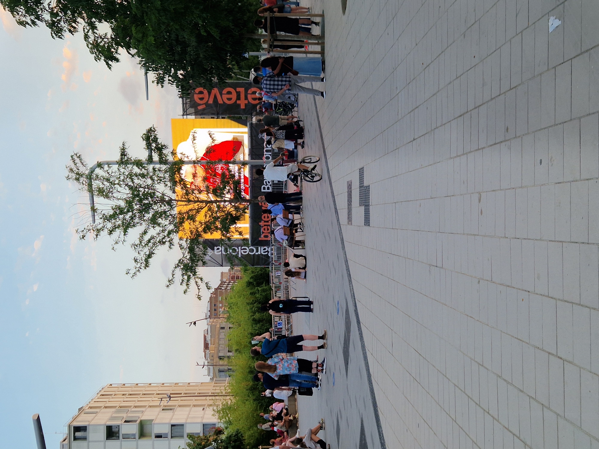 In Barcelona, Spain, a wide, paved plaza with a light gray, rectilinear tile pattern serves as the foreground. Several people are scattered across the plaza, some standing, some sitting on low walls or directly on the ground. A man in a white t-shirt and shorts is riding a bicycle towards the right side of the frame. To the left, a modern, beige building with multiple windows dominates the background. In the center, a large, illuminated outdoor screen displays a red graphic with white text that appears to read "BETEF" and "BARCELONA" vertically. Adjacent to the screen, a tall, dark banner with orange lettering, partially obscured by trees, displays "eTeve". Lush green trees and foliage are visible throughout the scene, with a bright sky overhead. Several streetlights are present, and a temporary barrier separates some of the seated individuals from the main plaza area.