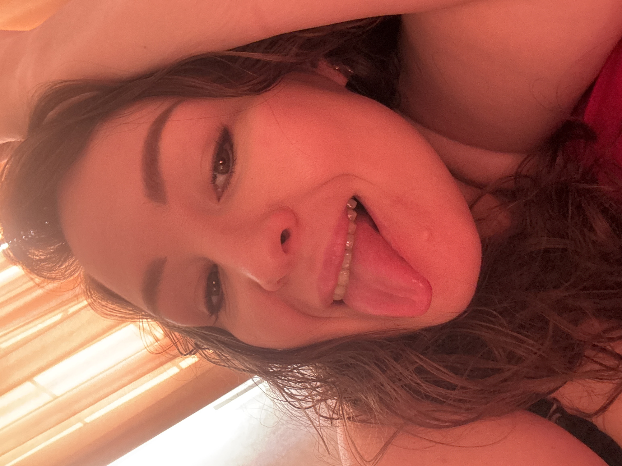 An upward-facing, close-up shot captures a young woman from Yuzhny, Russia, lying down and looking directly at the camera. Her head is tilted, giving the impression of an inverted perspective. She has long, wavy dark brown hair framing her face and extending around her. Her brown eyes are open, with visible eyeliner or mascara. A playful expression is conveyed by her tongue sticking out, which features a prominent piercing adorned with several small, white, beaded elements. Her skin has a warm, reddish hue, likely influenced by the lighting or a filter. A hint of a red garment, possibly a top or bra, is visible on her upper right side. In the background, what appears to be horizontal blinds or curtains are visible in the top left, suggesting an indoor setting near a window. The overall lighting is soft and warm. No text is visible in the image.