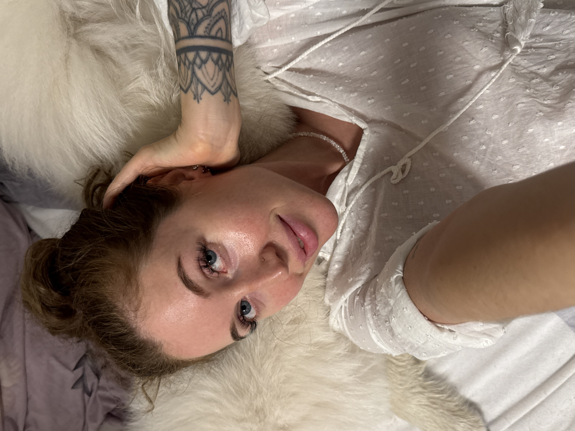A fair-skinned woman with light brown hair and blue eyes lies on her back. She wears a white blouse with small dots. A tattoo adorns her left forearm. She rests on a soft white fur-like material. The setting appears to be a bed, with a muted purple bedsheet visible. The image was likely taken in an indoor setting. It is not possible to verify the location in Chaikovsky, Russia.