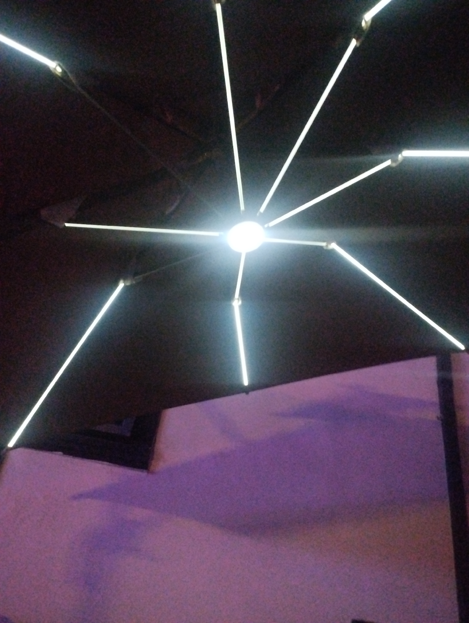 A low-angle shot captures a ceiling-mounted light fixture radiating light beams outwards. The fixture is positioned in the upper center of the frame, and its light reflects off the ceiling. Below the fixture, a wall with a textured surface is visible, illuminated with a purple hue. A dark object or shadow appears near the wall's edge. The location is Tumfure, Nigeria.