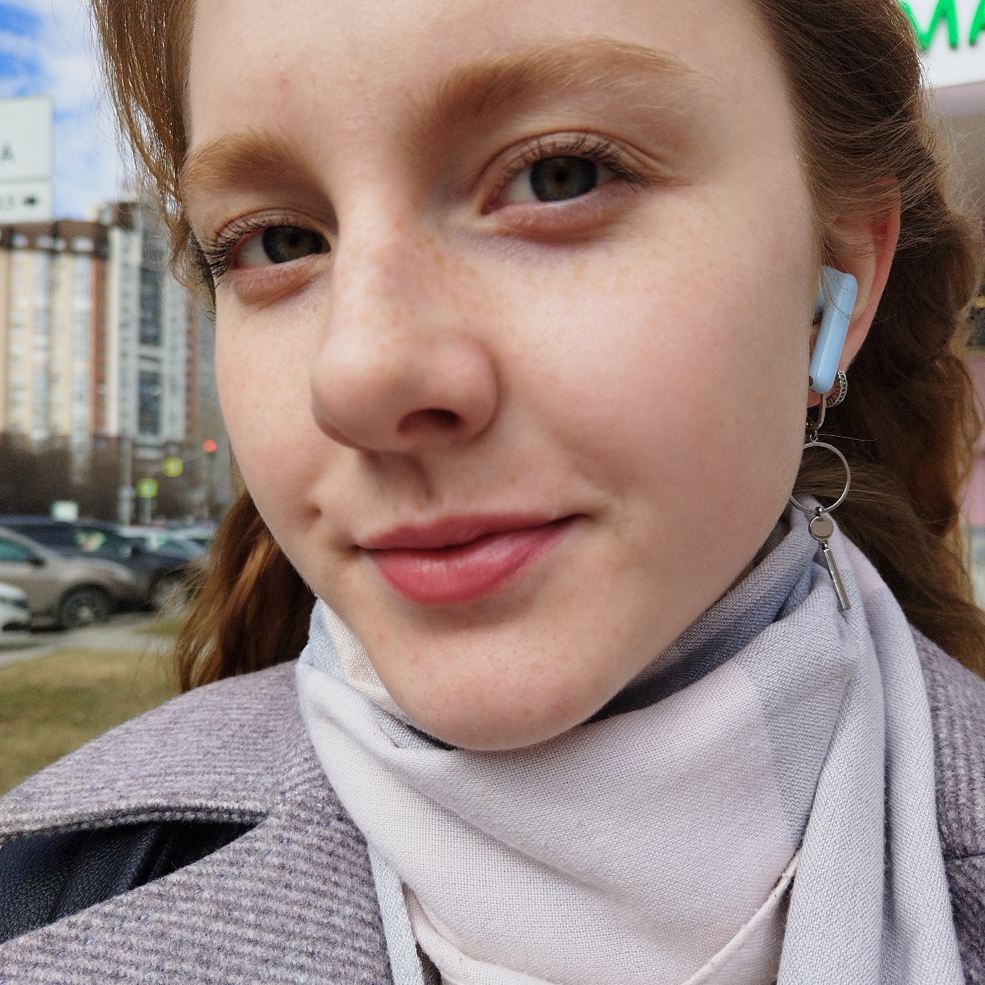 A close-up outdoor portrait features a young woman with fair skin, light reddish-brown hair, and numerous freckles across her nose and cheeks. She has green or hazel eyes and looks directly at the viewer with a subtle smile. Her hair is partially swept back, and strands frame her face.
She wears a light blue earbud in her right ear, along with a silver-toned dangle earring consisting of a large hoop and a small, elongated cylindrical pendant. A light gray or off-white scarf with a textured pattern is wrapped around her neck. She is dressed in a light gray or lavender-colored coat, possibly with a subtle herringbone weave and sparkling threads, worn over a dark garment visible at the lapel.
The background, out of focus, reveals an urban setting with multi-story buildings, indicating a city or town street. Several parked cars line what appears to be a road or sidewalk. A red traffic light is visible in the mid-ground. The lighting suggests it is daytime, and the presence of a coat and scarf implies cooler weather.
Faint text is visible in the background: the letters "MA" appear on a green sign in the upper right, and a partial "A" is on a blurry sign in the upper left. The overall scene captures a casual moment in an everyday city environment.