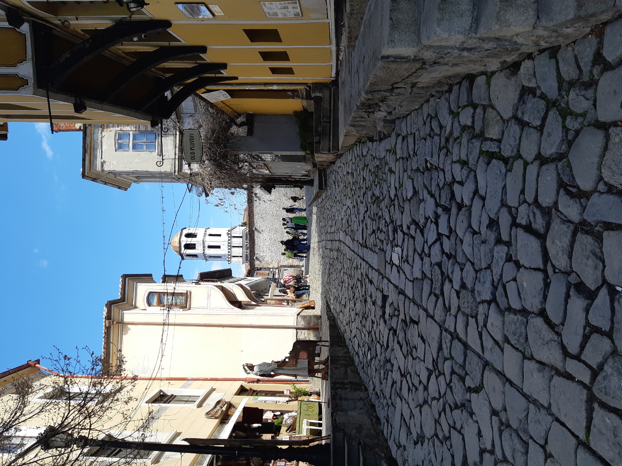 A street scene in Plovdiv, Bulgaria, features a cobblestone road running from the foreground to the background. The road is flanked by buildings on both sides, with a mix of yellow and white facades. A notable feature is the tower in the background, with a golden dome. Several people are visible in the distance. A sign reading "Old Plovdiv" is attached to a building on the left side. A black awning is present at the top left of the frame.