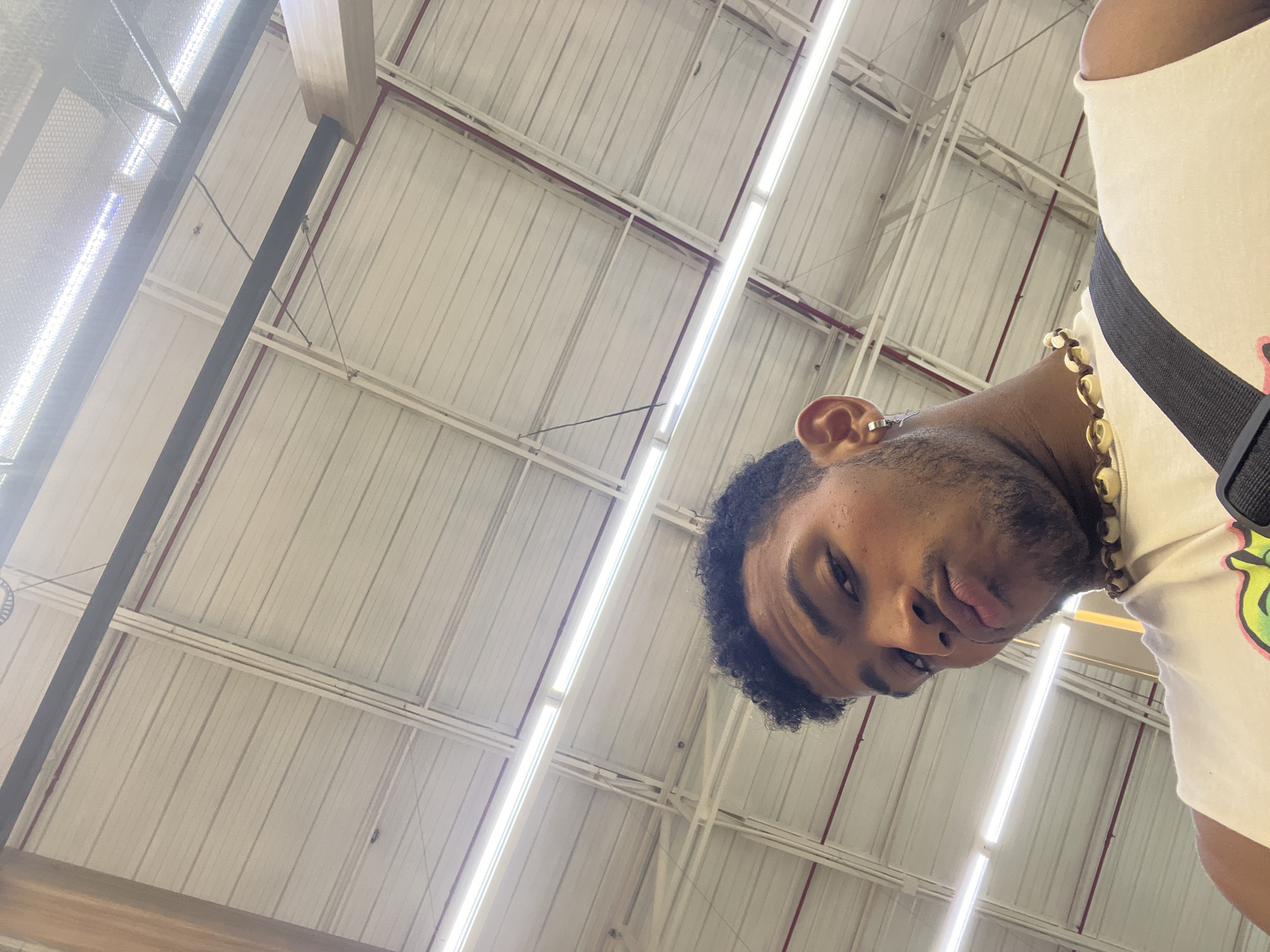 An upward-angle shot shows a man with curly black hair, a gold chain necklace, and an earring in his left ear. He is wearing a white t-shirt with a black strap or belt across his chest. The background consists of a white, corrugated metal ceiling with visible support beams and fluorescent lighting. The location is not specified in the information provided. The city of Porlamar, Venezuela is not evident from the image.
