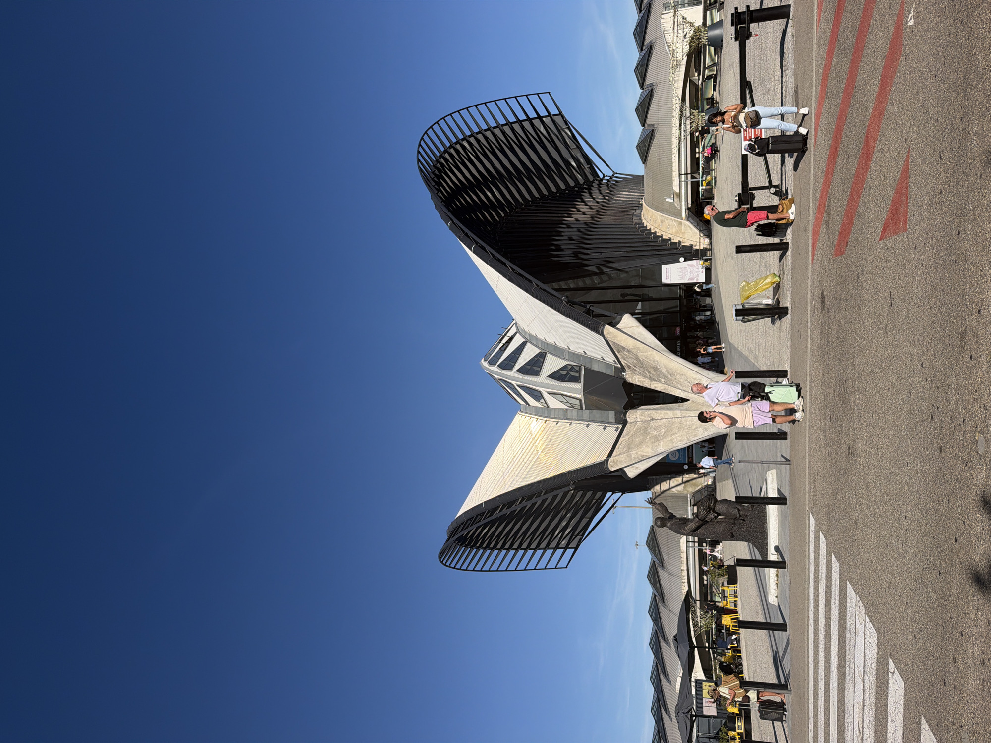 The image captures an outdoor scene in Colombier-Saugnieu, France, likely at an airport or a similar transportation hub, given the presence of a pedestrian crossing and people with luggage. The setting is modern and architectural, featuring a distinctive building with a curved, metallic roof structure that has a ribbed appearance. The sky is a clear, deep blue, indicating fair weather and likely midday with bright sunlight, casting sharp shadows.
Several individuals are visible. Two people stand near a trolley suitcase, one appearing to be a woman with dark hair wearing a white top and light-colored pants, and the other a man in a dark t-shirt and red shorts. Further down, two other individuals, a man and a woman, are seen near a suitcase, with the woman wearing a white top and light-colored pants, and the man in a light-colored t-shirt and light shorts. Another person is visible closer to the building, with a yellow bag. In the background, there are tables and chairs under umbrellas, suggesting an outdoor seating area for a cafe or restaurant.
The architecture is a prominent feature, with its dramatic, wing-like curves and geometric windows. The ground is asphalt, marked with red lines and a white pedestrian crossing. The overall mood seems relaxed and observational, with people appearing to be waiting or traveling.
There is some visible text, including a sign on the building that appears to read "AEROPORT DE LYON" and "L'EUROPE N'EST PAS LOIN". Another sign closer to the people has some illegible text. The presence of an airport name suggests the location is indeed Lyon Airport.
