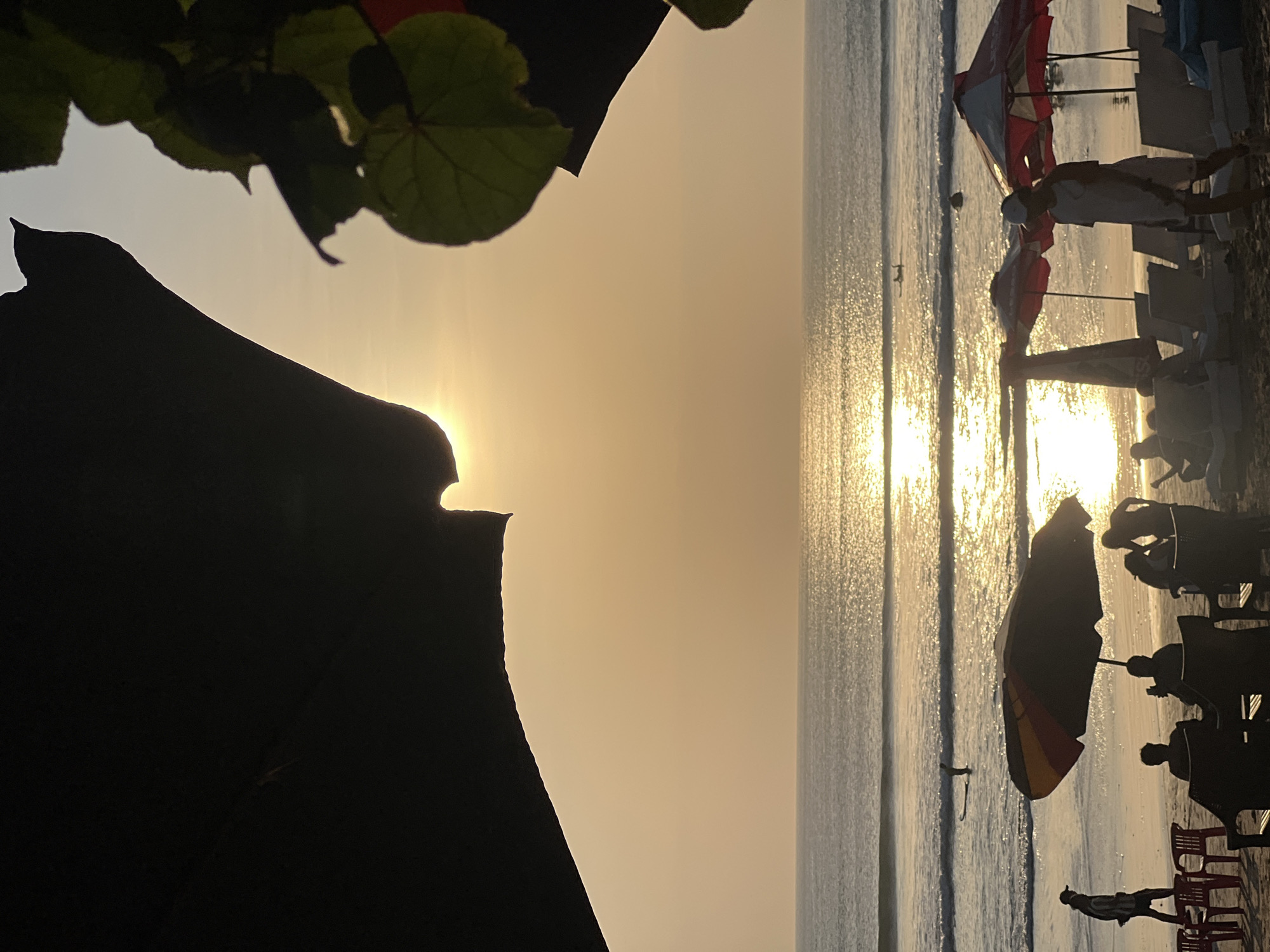 The image depicts a sunset scene at Kuta Beach in Indonesia. The sun is low on the horizon, casting a warm, golden glow across the sky and the ocean. The water is calm with gentle waves, and the sun's reflection creates a shimmering path across its surface.
In the foreground, beach umbrellas and lounge chairs are set up on the sand, suggesting a relaxed atmosphere. Several people are visible, some sitting under umbrellas, while others are walking along the shore or in the water. One person is seen walking towards the left of the frame, while others are silhouetted against the bright sky. In the ocean, there are a few individuals on surfboards, enjoying the waves.
The image is framed by dark, silhouetted shapes in the upper left and upper right corners, which appear to be large umbrellas or natural foliage. These elements add depth and contrast to the scene, drawing attention to the luminous sunset over the ocean. The overall mood is serene and tranquil, characteristic of a beach at dusk.
