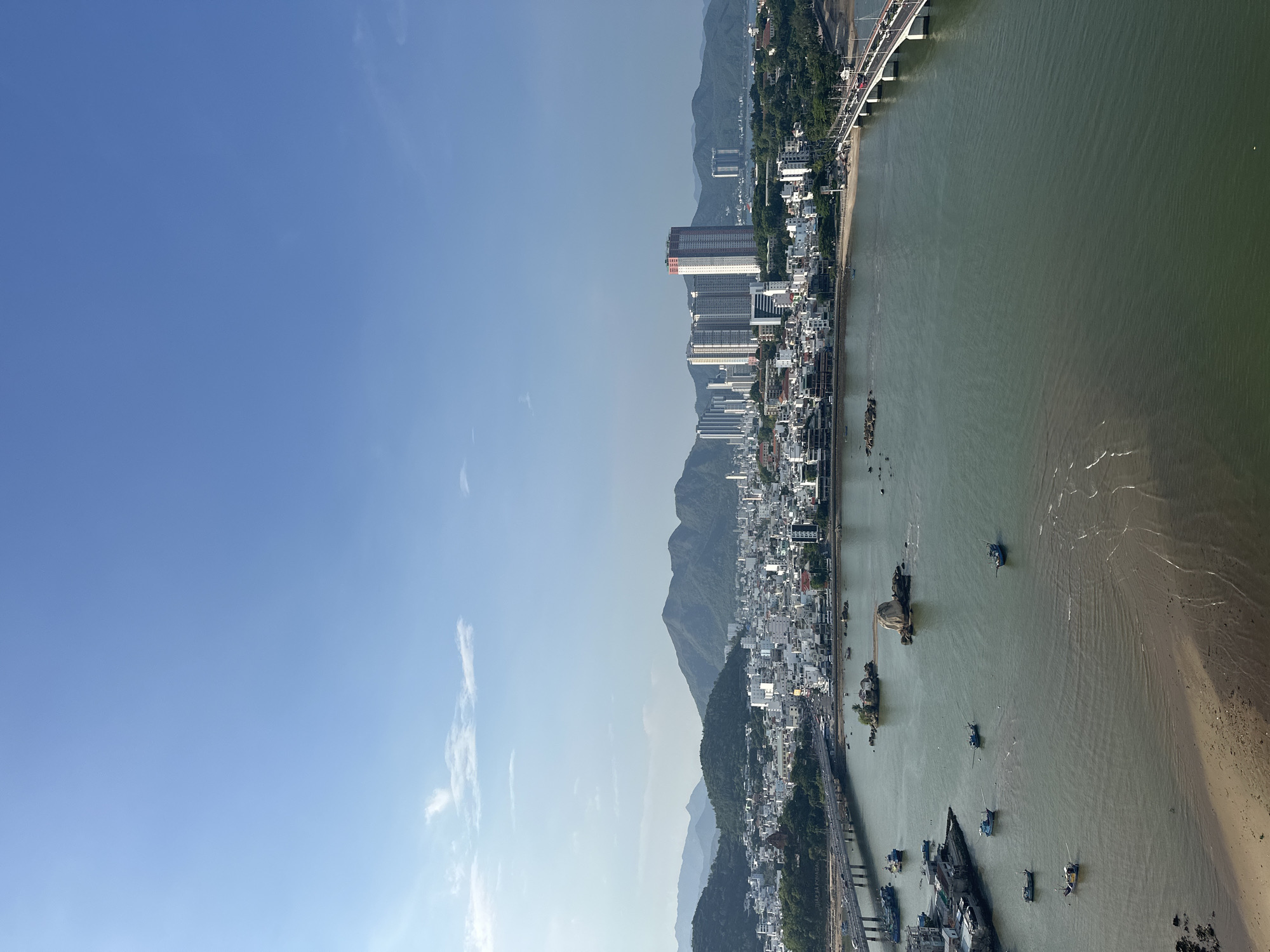 This is a high-angle, panoramic photograph of a coastal city, likely Nha Trang, Vietnam, on a clear day with a blue sky and scattered wispy clouds. The foreground features a body of calm, greenish-brown water, likely a bay or river, with a sandy shoreline visible on the left. Several small, dark-hulled fishing boats are scattered across the water, some with white cabins.
The middle ground showcases a densely populated urban area with a mix of white and light-colored buildings, ranging from low-rise structures to numerous multi-story apartment buildings and a few prominent high-rise towers. A large, distinctly shaped rock formation juts out of the water near the shore. A bridge with vehicles traversing it spans the water from the left side of the frame, connecting to the city. Another bridge with traffic is visible on the right side of the frame, also connecting to the city.
In the background, lush green mountains form a natural backdrop to the city. The overall impression is one of a vibrant coastal city nestled between the sea and mountains, with evidence of both urban development and natural scenery. The lighting suggests it is daytime, possibly late morning or early afternoon. No people are clearly discernible in the image.