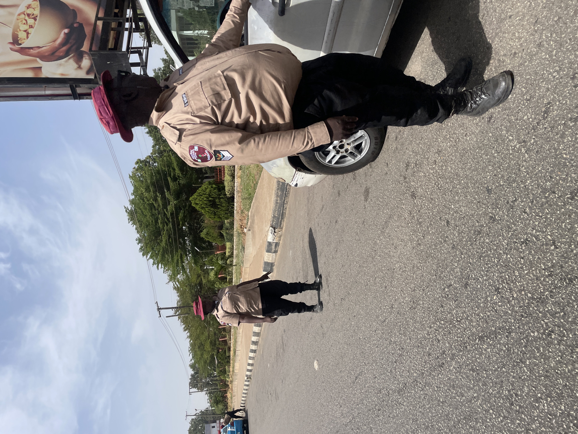 An adult male wearing a khaki shirt, black pants, black boots, and a red hat leans out of a white vehicle. The man is wearing a badge on his left shoulder, and his right hand rests on the vehicle's front tire. The scene takes place on a paved road with trees and telephone poles visible in the background. A blue and white car is driving on the opposite side of the road, and an advertisement is to the upper left of the man.