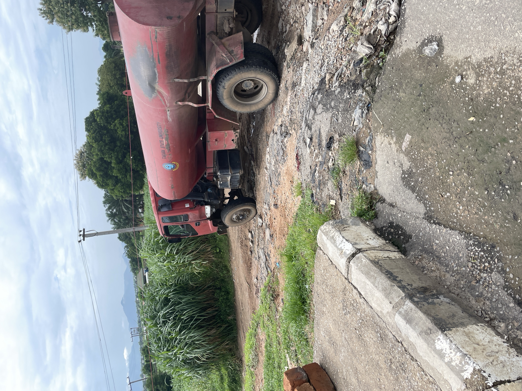 The image shows a red tanker truck parked on a dirt road next to a paved area. The truck has a large cylindrical tank on its chassis and a visible logo and text on the side of the tank. The text reads "TARABA STATE UNIVERSITY" along with some unreadable text and a crest. The truck appears to be weathered, with some rust and dirt visible on its surface.
The scene is set in a semi-rural or urban fringe environment, with lush green vegetation, including tall grasses and trees, in the background. In the distance, mountains are visible under a cloudy sky. Power lines and utility poles are also present, suggesting the presence of infrastructure. The paved area next to the dirt road has some puddles of water and debris scattered around, indicating potential drainage issues or recent rainfall. Some bricks are stacked in the foreground, possibly left from construction or repair work.
The time of day appears to be daytime, with natural light illuminating the scene. The weather seems overcast, with a cloudy sky, but not actively raining. There are no people visible in the image. The overall impression is one of a working vehicle in a somewhat unkempt or developing area. The presence of the university's name on the truck might suggest it is used for water supply or other services to the institution. The context provided indicates the location is Jalingo, Nigeria.