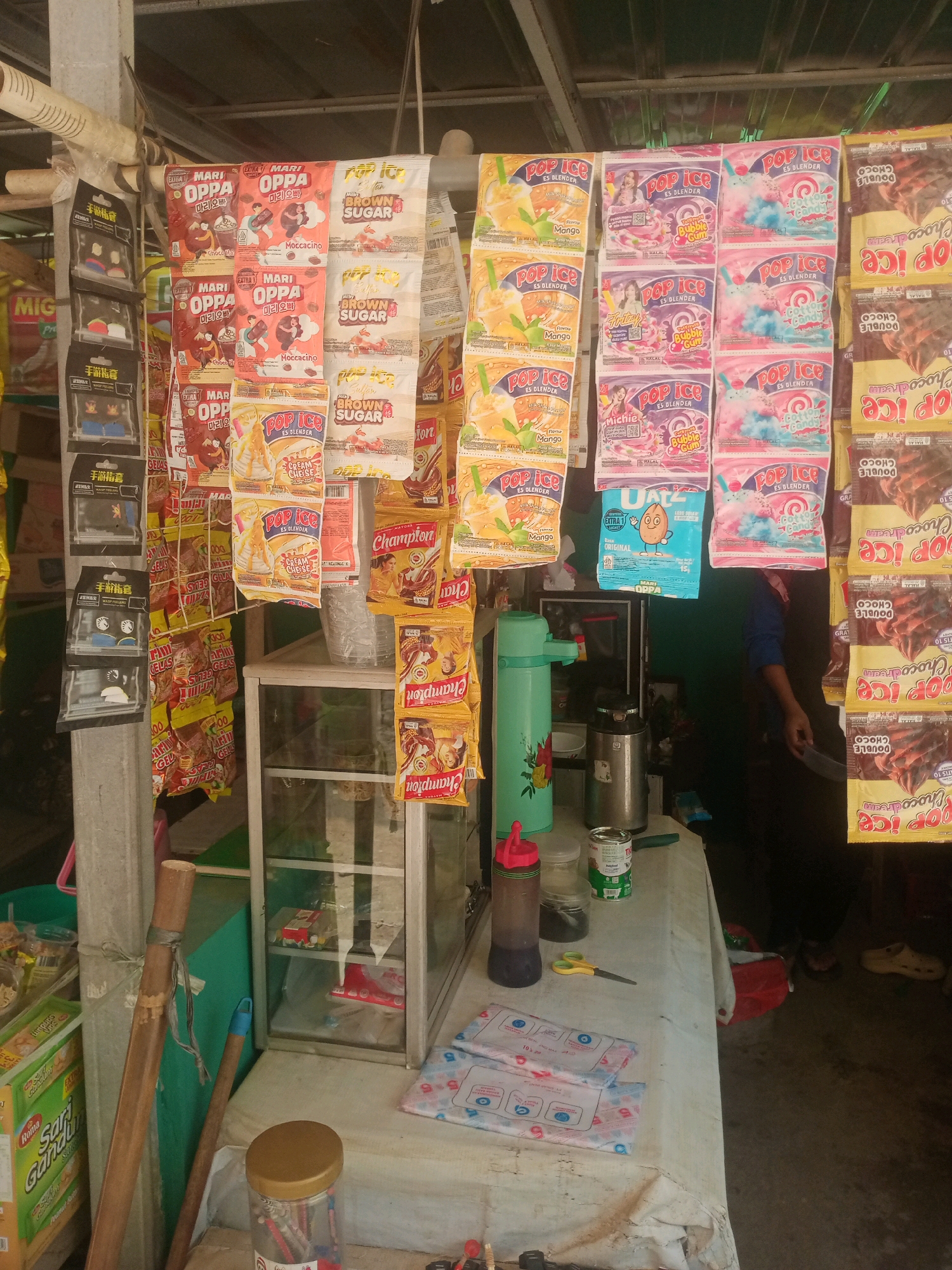 A roadside stall selling various food and drink items is depicted. Products are hung from a wooden beam, creating a cluttered but organized display. On the left, several small, dark-colored packages with Chinese writing are arranged vertically. Adjacent to these are rows of larger packets, some featuring the "Mari Oppa" branding with cartoonish figures and Korean text, and others with "Pop Ice" branding showcasing various flavors like "Brown Sugar," "Cream Cheese," and "Cotton Candy." Further to the right, a stack of "Champion" brand packets and "Pop Ice Double Choco Dream" packets are visible.
In the center of the image, a small glass display cabinet contains some boxed items. To the left of the cabinet, a tall, teal-colored thermos sits on a table covered with a light-colored cloth. Several cans and bottles, including what appears to be a can of condensed milk and a bottle with a red lid, are also on the table. A pair of yellow scissors rests on the cloth near the thermos.
The background is indistinct, with hints of a green wall and a dark interior. A person's arm and shoulder are visible on the far right, suggesting an attendant or customer. The ground appears to be a dirt or concrete surface. Two wooden poles are propped against the stall, one longer and thicker than the other, and a blue handle is attached to the shorter pole. A clear plastic container filled with colorful pencils is on the table in the foreground.