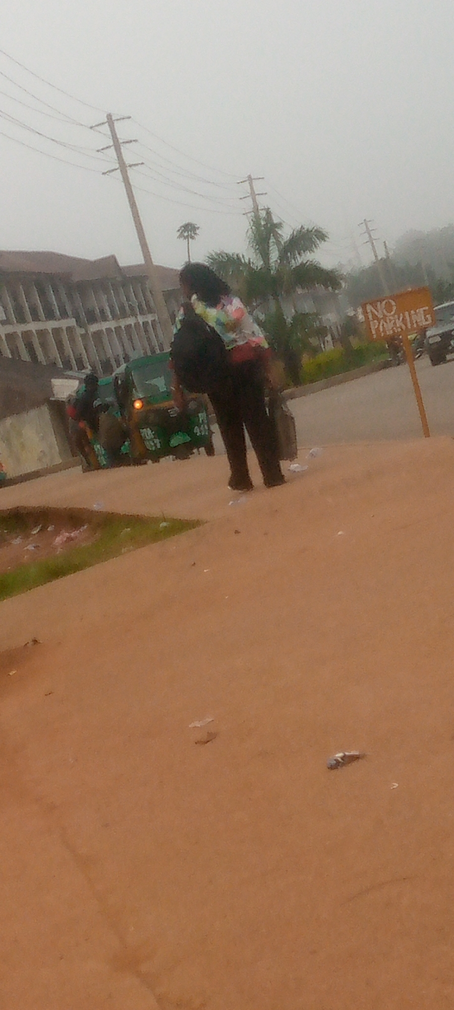 The image depicts an outdoor scene on a hazy day, possibly in Gura Top, Nigeria. In the foreground, a person wearing dark pants and a colorful jacket with a backpack is standing. Their back is to the camera. Behind them, a green auto-rickshaw is visible. Further in the background, there is a building with multiple floors and a faded facade. To the right of the auto-rickshaw, a wooden sign with the words "NO PARKING" is planted in the ground. Utility poles with wires are also present in the background. The ground is a reddish-brown dirt or sand color, with scattered debris.