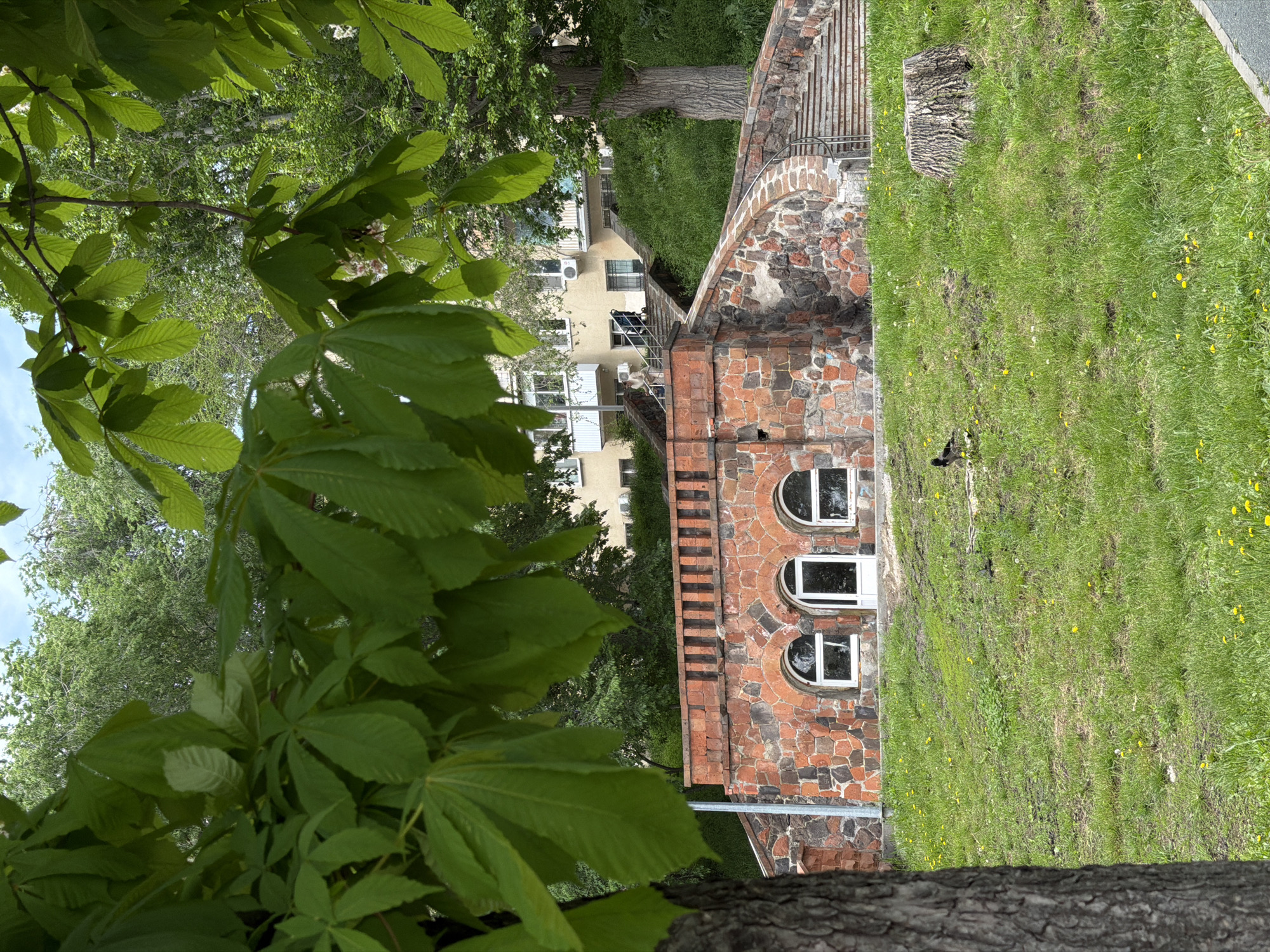 A color photograph of a building is framed by a tree in the foreground. The building appears to be constructed with red and brown stones, and has three arched windows in a vertical row. A set of stairs runs along the side of the building. Grass covers the ground to the right of the building.
In the background, another building can be seen, with multiple windows and air conditioning units visible. Additional trees and foliage fill the background. The photograph appears to have been taken outdoors in a green, urban environment.
The image depicts a building near the Volga River in Saratov, Russia.