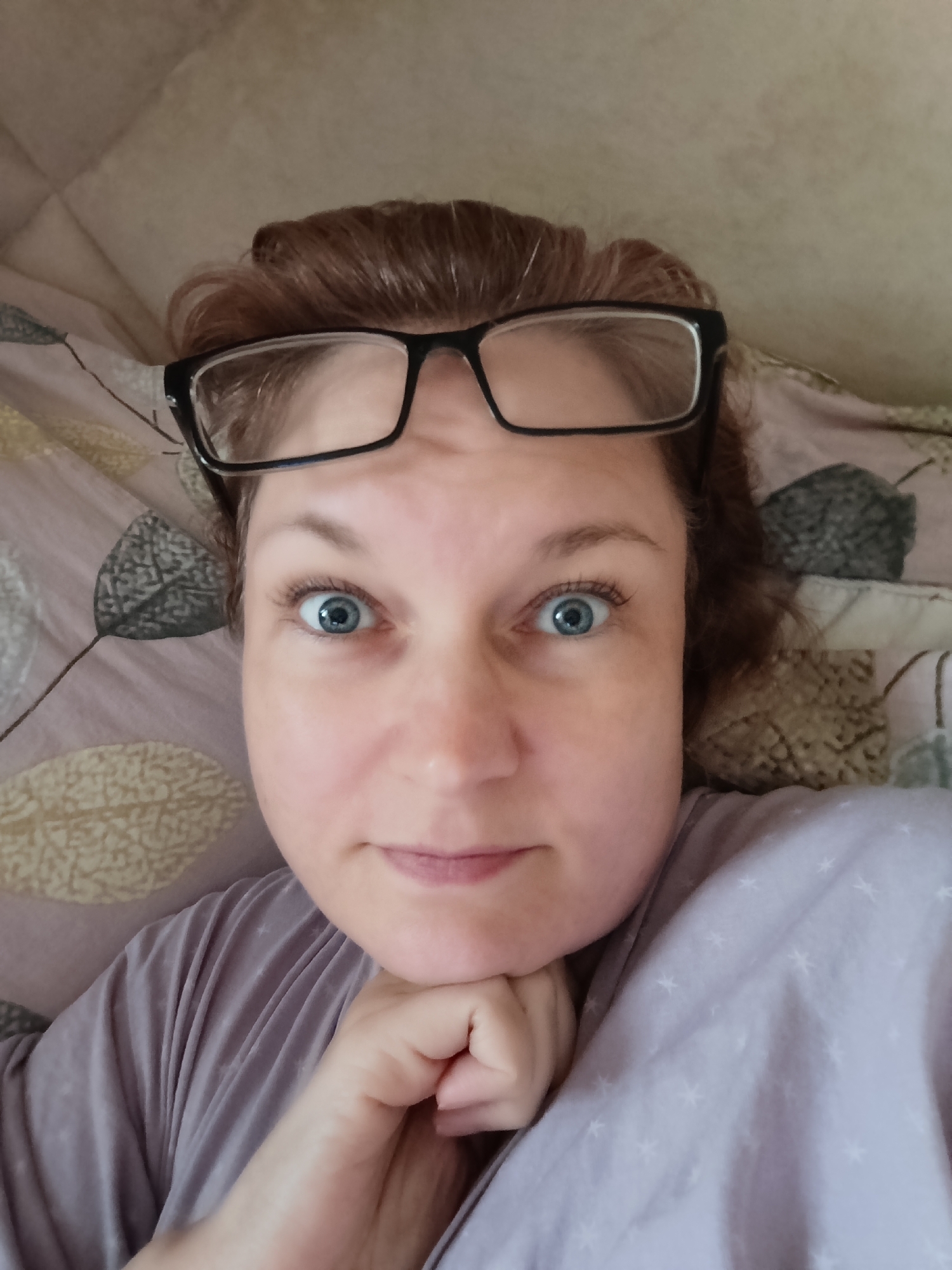 A close-up, slightly tilted, selfie shows a woman with reddish-brown hair and blue eyes. She is wearing black-rimmed glasses and a light purple pajama top with a subtle star pattern. Her hands are clasped and resting under her chin, supporting her head. The background appears to be a bed with a patterned comforter, featuring leaf-like designs in muted tones. The lighting suggests it is daytime, but there are no external windows or cues to indicate the weather. The woman's expression is one of surprise or wide-eyed curiosity. The image was likely taken indoors, in a home setting in Марковский, Russia.