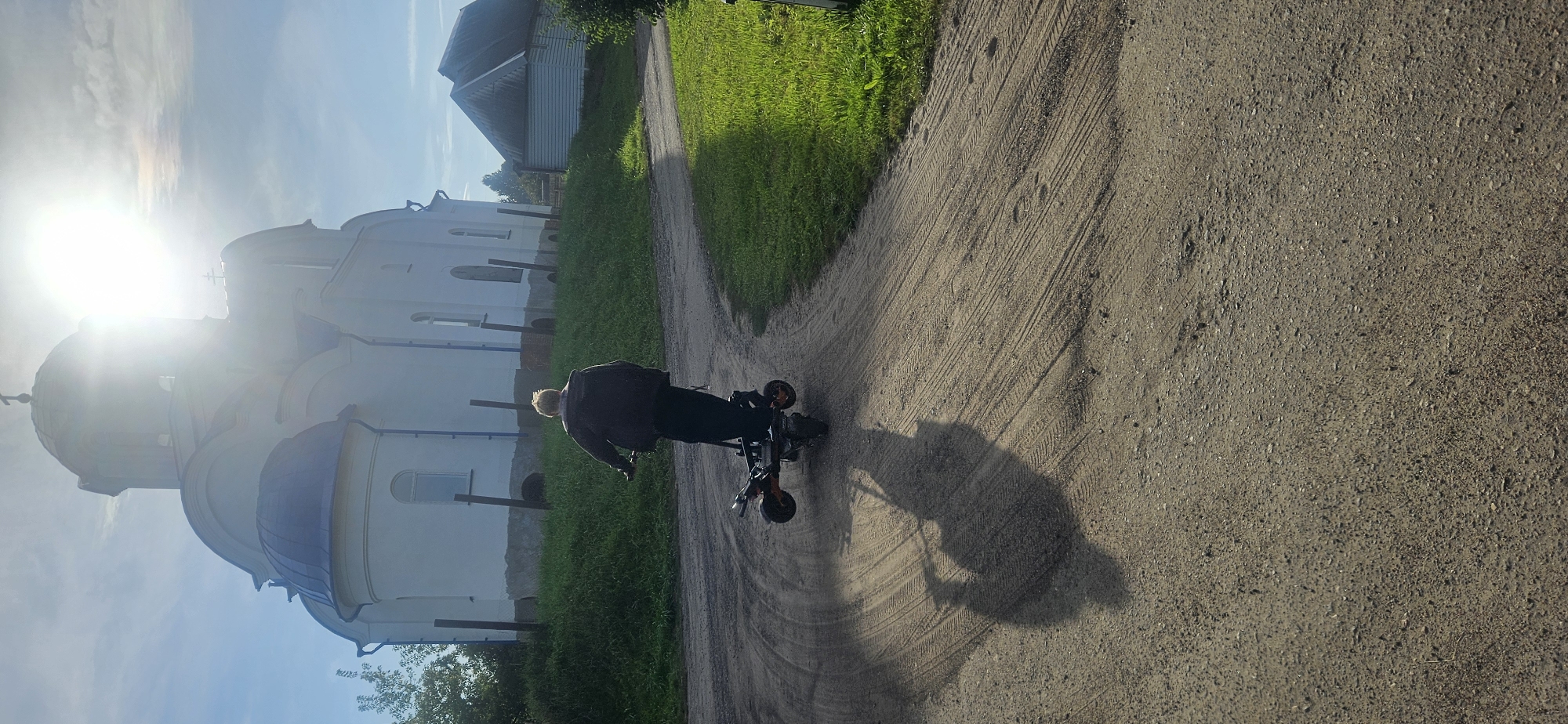 A person with blond hair is riding a three-wheeled electric scooter on a dirt road. The scooter has an orange frame and black handlebars. The person is wearing a dark jacket and dark pants. They are facing away from the camera, riding towards a white building with a blue dome, which appears to be a church. The sun is shining brightly in the sky, causing a lens flare and casting a long shadow of the person and scooter on the road. Green grass is visible on the sides of the road and around the building. A smaller building with a dark roof is visible in the background to the right of the church. The overall scene suggests a rural or suburban setting in Vitsebsk, Belarus.