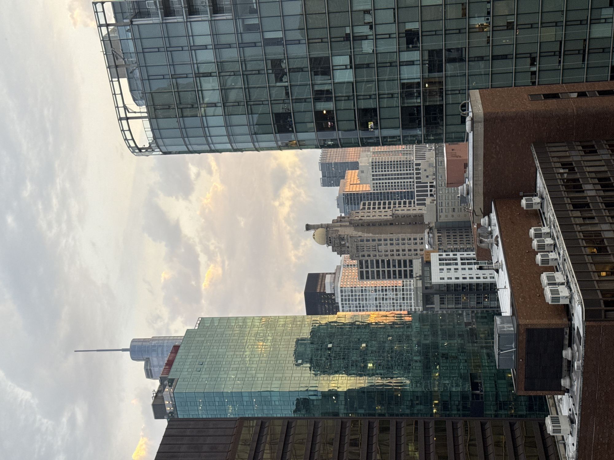 The image shows a cityscape with numerous skyscrapers in Chicago, Illinois, United States of America. The photograph captures the buildings from a slightly elevated perspective, offering a view of their glass facades and architectural details. The sky is filled with clouds, with hints of sunlight breaking through, casting a soft glow on the buildings. In the foreground, a few of the buildings' rooftops are visible, featuring air conditioning units and other mechanical equipment. The overall impression is of a busy urban environment bathed in the muted light of a cloudy day.