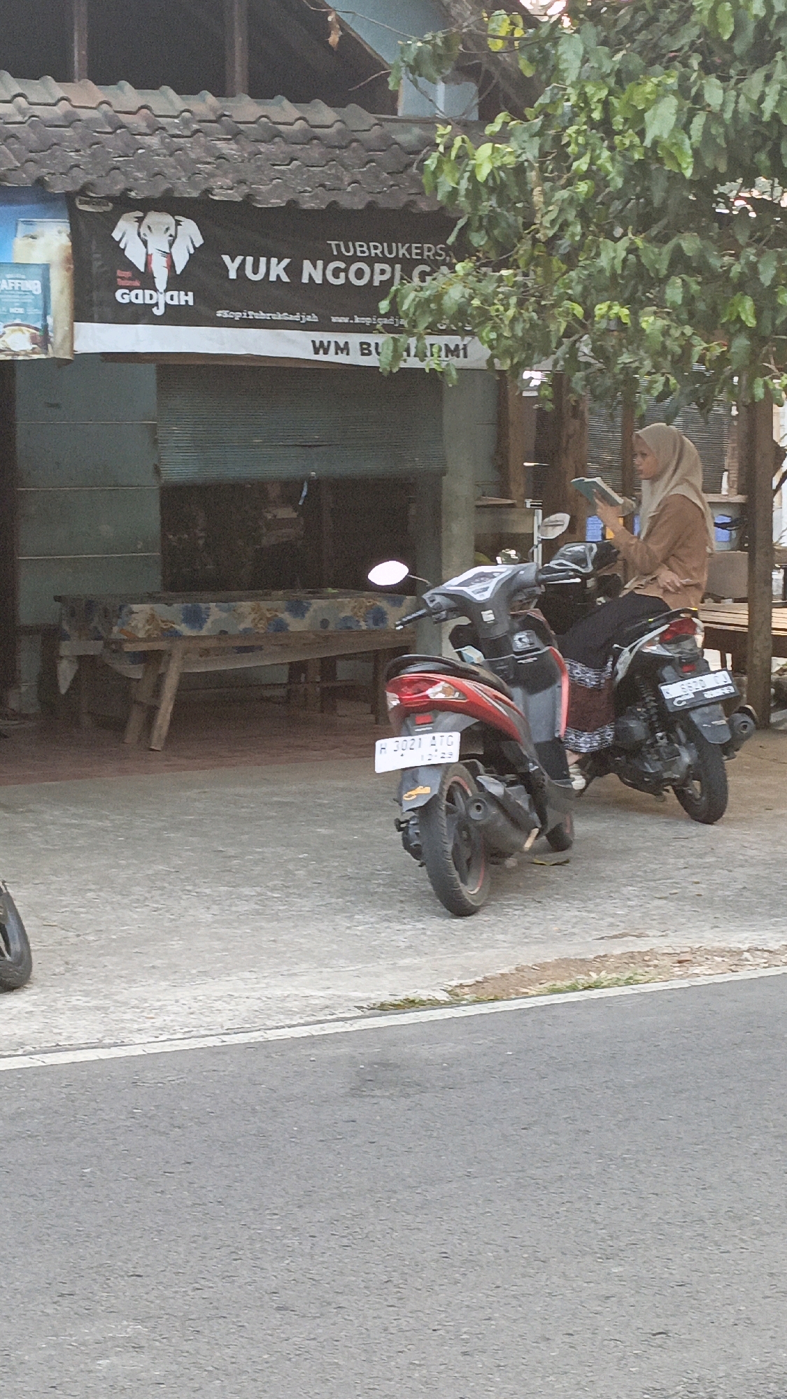 An outdoor scene depicts a small roadside eatery or shop in Salatiga, Indonesia, with two motorcycles parked in front of it. A black banner with white text and an elephant graphic is displayed above the shop entrance. The text on the banner reads "TU BRUKERS, YUK NGOPIGAJAH" and includes a hashtag "#kopitubrukGadjah" and a website address "www.kopigadjah.com". Below this, another line of text reads "WM BUMIRMI".
A woman wearing a hijab and a brown top is seated on one of the motorcycles, engrossed in a book or tablet she is holding. The motorcycles are parked on a concrete surface adjacent to a paved road. In the background, a long, wooden table covered with a patterned cloth is visible. Trees with green foliage are present in the upper right portion of the image, providing some shade. The overall atmosphere suggests a casual, everyday scene in a localized commercial area.