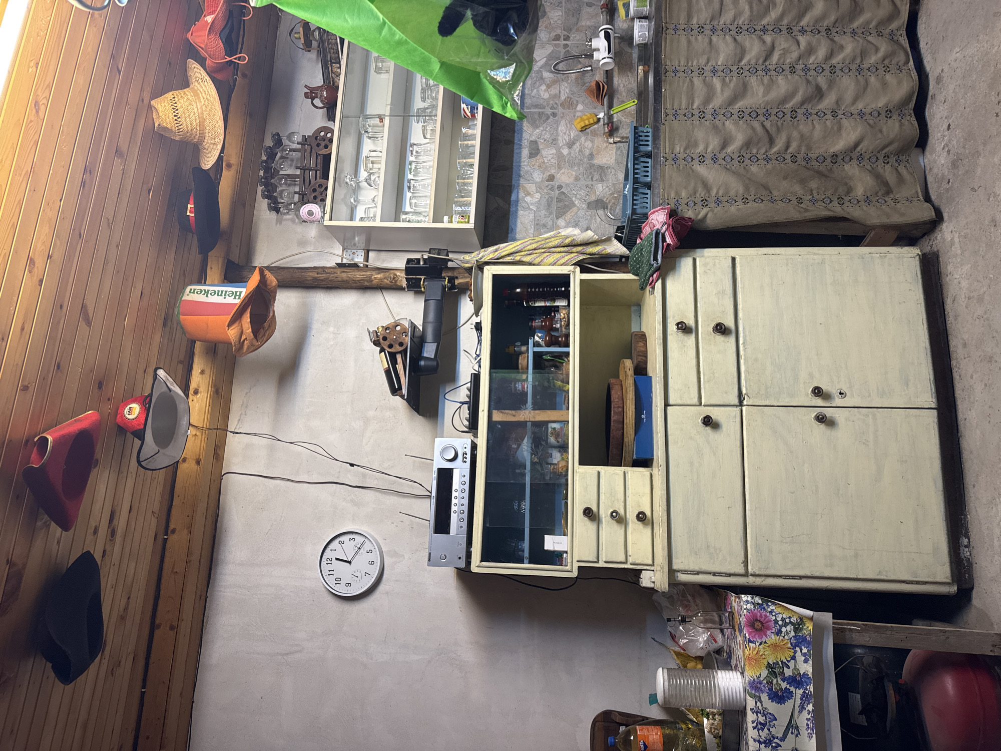 This appears to be a cluttered interior shot of a room, possibly in a home or storage area. The scene is a bit messy and has a rustic or lived-in feel.
**Key elements:**
* **Furniture:** There is a large, older cabinet with several doors. Next to it stands a smaller open cabinet with glass display cases.
* **Objects:** The room is filled with various objects including bottles, glasses, hats hanging on a wooden wall, tools, and electronic equipment like a sound system. A clock is on the wall, reading approximately 10:08.
* **Environment:** The walls are a mix of wood paneling and plastered surfaces. The floor appears to be a mix of concrete.
* **People and Actions:** The image does not show any people or ongoing actions.
Given that the room contains a "Heineken" themed item and a "FANTA" bottle, coupled with a slightly disorganized aesthetic, the setting is most likely a storage area or a room inside a home that has not been tidied.
While it is not confirmed where the photo was taken, there is a mention of Blajievo, Bulgaria, on the Heineken box.