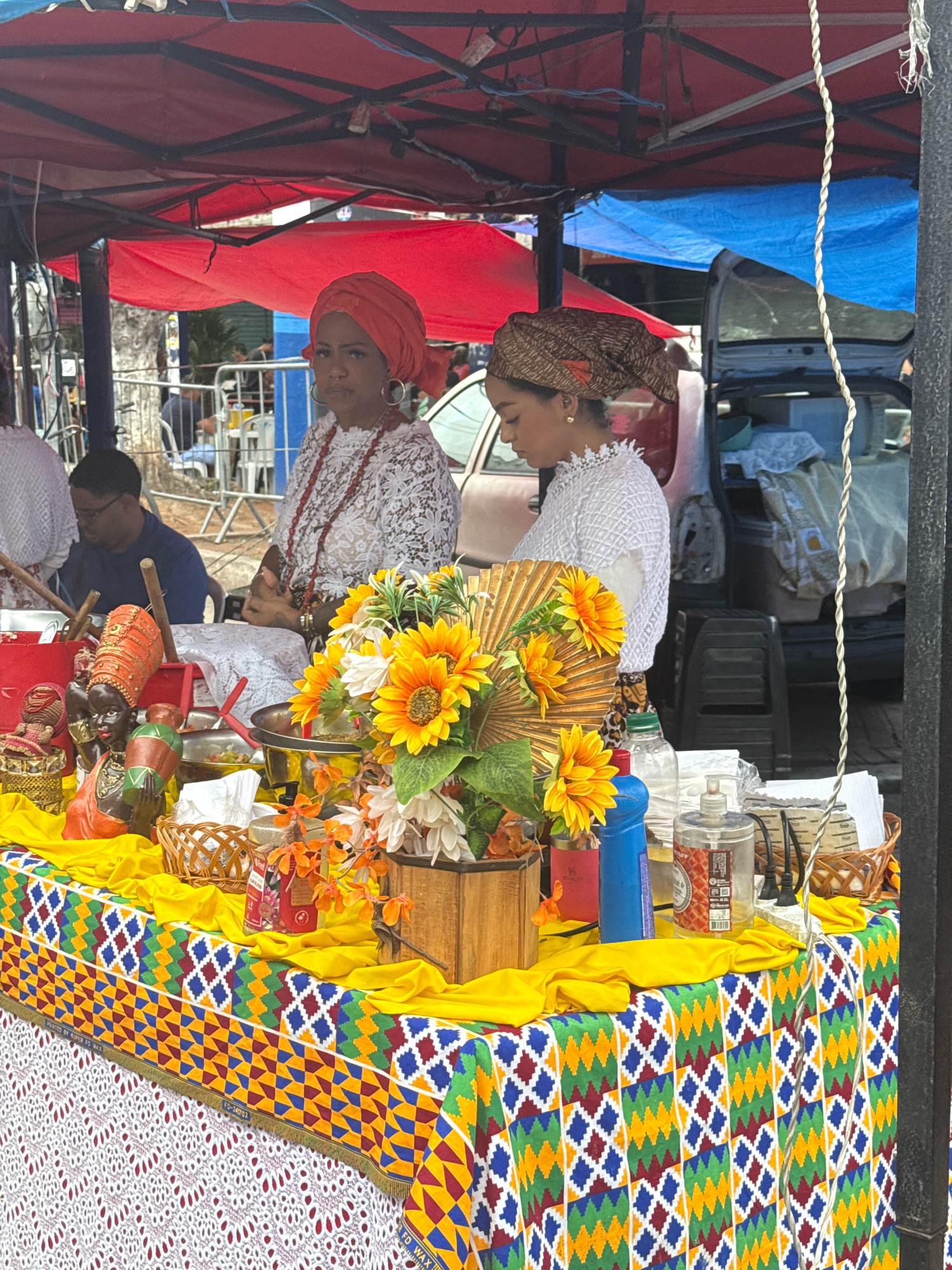 An outdoor market stall is set up under a red and blue canopy. Two women are visible behind the stall. The woman on the left wears a white lace top and a bright orange headscarf. She has a large circular earring and a necklace made of red beads. The woman on the right wears a white top with an intricate pattern and a brown patterned headscarf. The stall is draped with a yellow cloth and a colorful fabric with a geometric pattern. On the stall, there are various items including several small decorative figurines, a fan made of straw with painted sunflowers, a wooden box, and bottles of cleaning products and hand sanitizer. The background shows a car with its trunk open and other market activities. The location is the city of Nilópolis, Brazil.