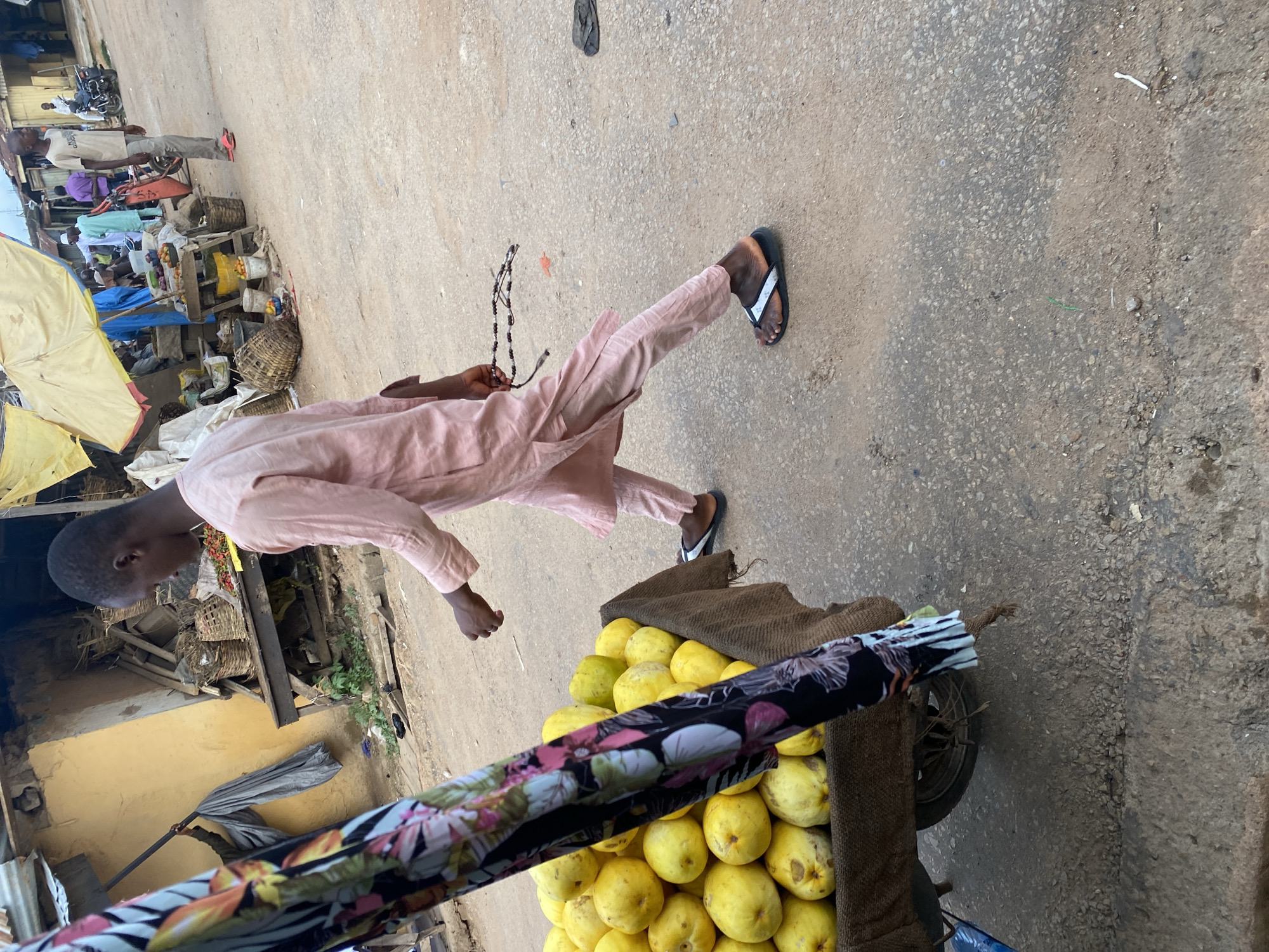 A young Black male in a light pink, loose-fitting traditional outfit walks from right to left, carrying a string of dark beads. He is wearing black flip-flops. To his left, a cart is filled with large yellow fruits, possibly guavas, covered with burlap fabric. The cart is partially obscured by a colorful fabric panel with a floral pattern. The scene appears to be a marketplace in Jos, Nigeria. In the background, there are market stalls with various goods, including baskets and what appears to be produce. Several other people are visible in the distance, engaged in market activities. The ground is paved with asphalt.