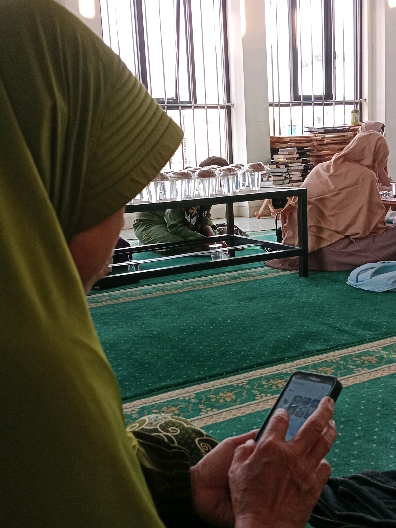 In the city of Purbayan, Indonesia, a group of individuals are gathered indoors, likely in a mosque or a similar community space, as indicated by the green carpet with a traditional pattern. In the foreground, a person wearing a green hijab is seated and holding a smartphone, their fingers interacting with the screen. Behind them, several other individuals are also seated on the carpet, some wearing hijabs and traditional Islamic clothing. A black metal table with several glasses of water is visible, as well as stacks of books and religious texts in the background. Large windows with vertical blinds line the back wall, allowing natural light to enter the space. The overall atmosphere appears to be one of quiet contemplation or religious observance.