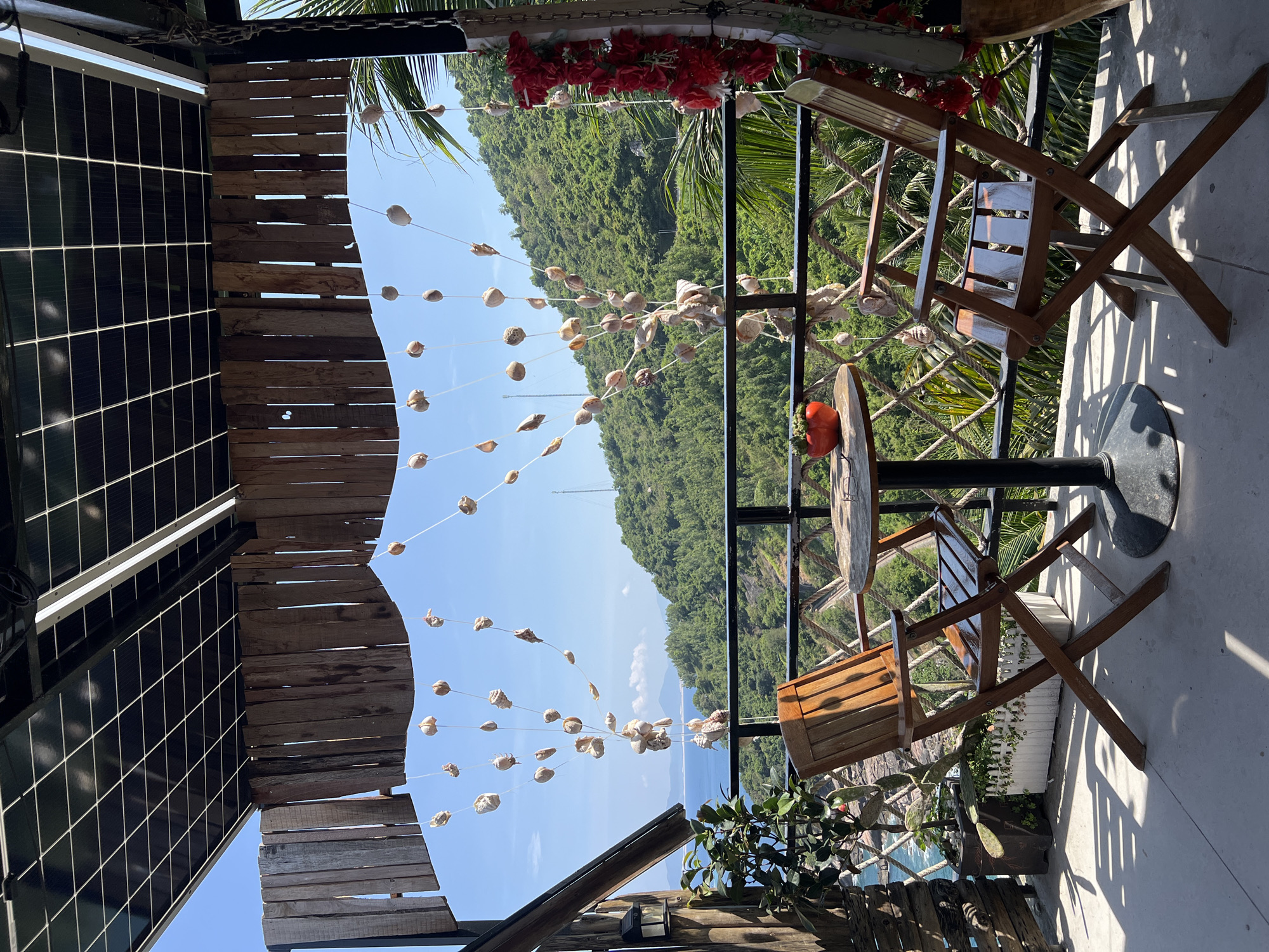 This is an outdoor scene in Hòa Xuân, Vietnam. A wooden platform and partial roof structure are on the left, with decorations including strung shells. To the right, there is a small round table with two wooden chairs. A railing divides the seating area from a view of lush, green mountains and a body of water in the distance. There are some decorative plants near the railing. The sky is clear blue.