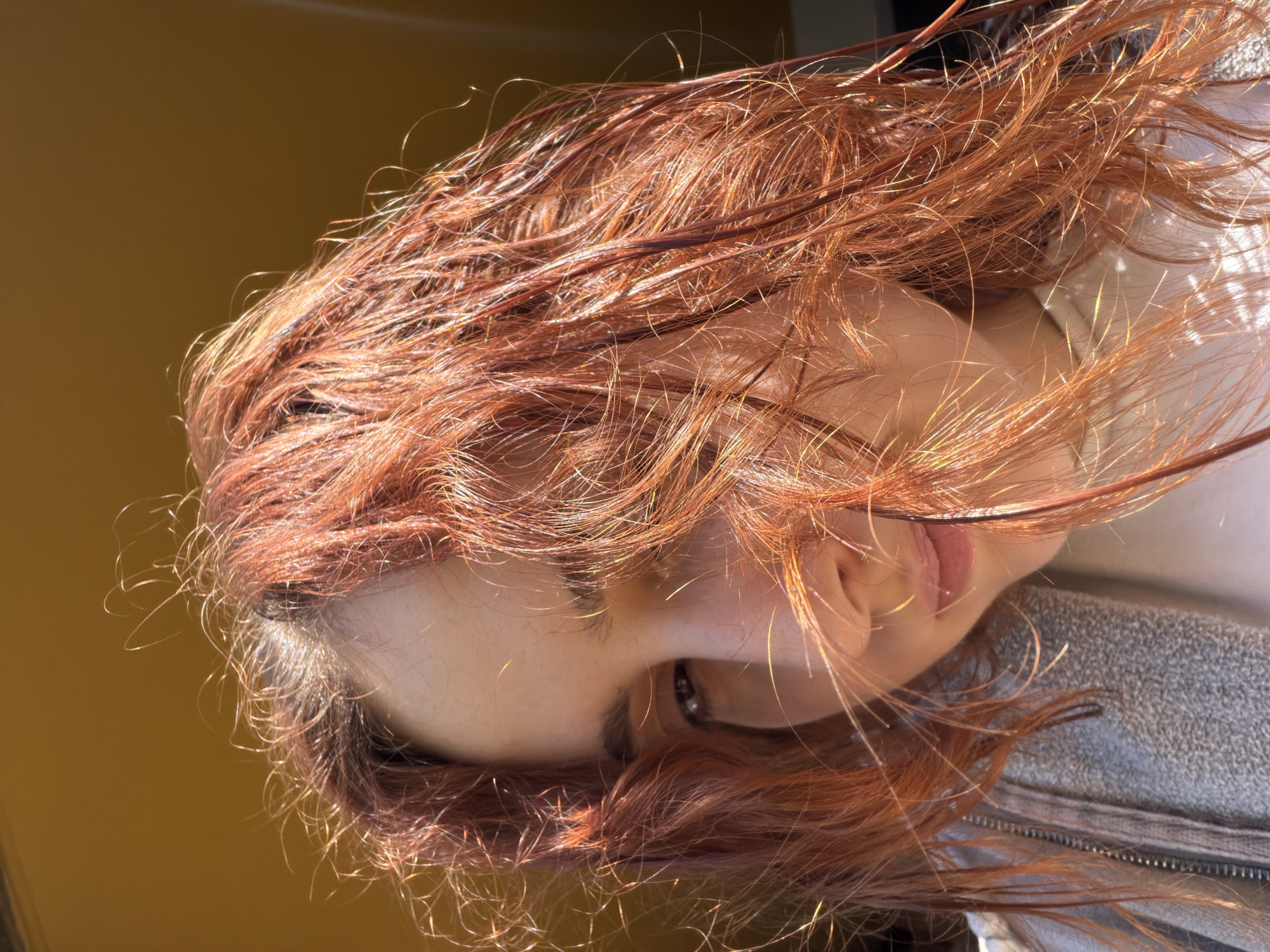 An upside-down, close-up shot focuses on a female face with fiery red hair. Her face is fair, and one eye is visible. The top of her face is oriented toward the left side of the frame, while the bottom half of her face angles towards the right. Wisps of red hair frame her face, which is set against a blurred background of what appears to be mustard yellow. The lower right portion of the image hints at the texture of a light grey garment. There are no immediate clues to ascertain whether it was taken in Strasbourg, France, or any other specific location.