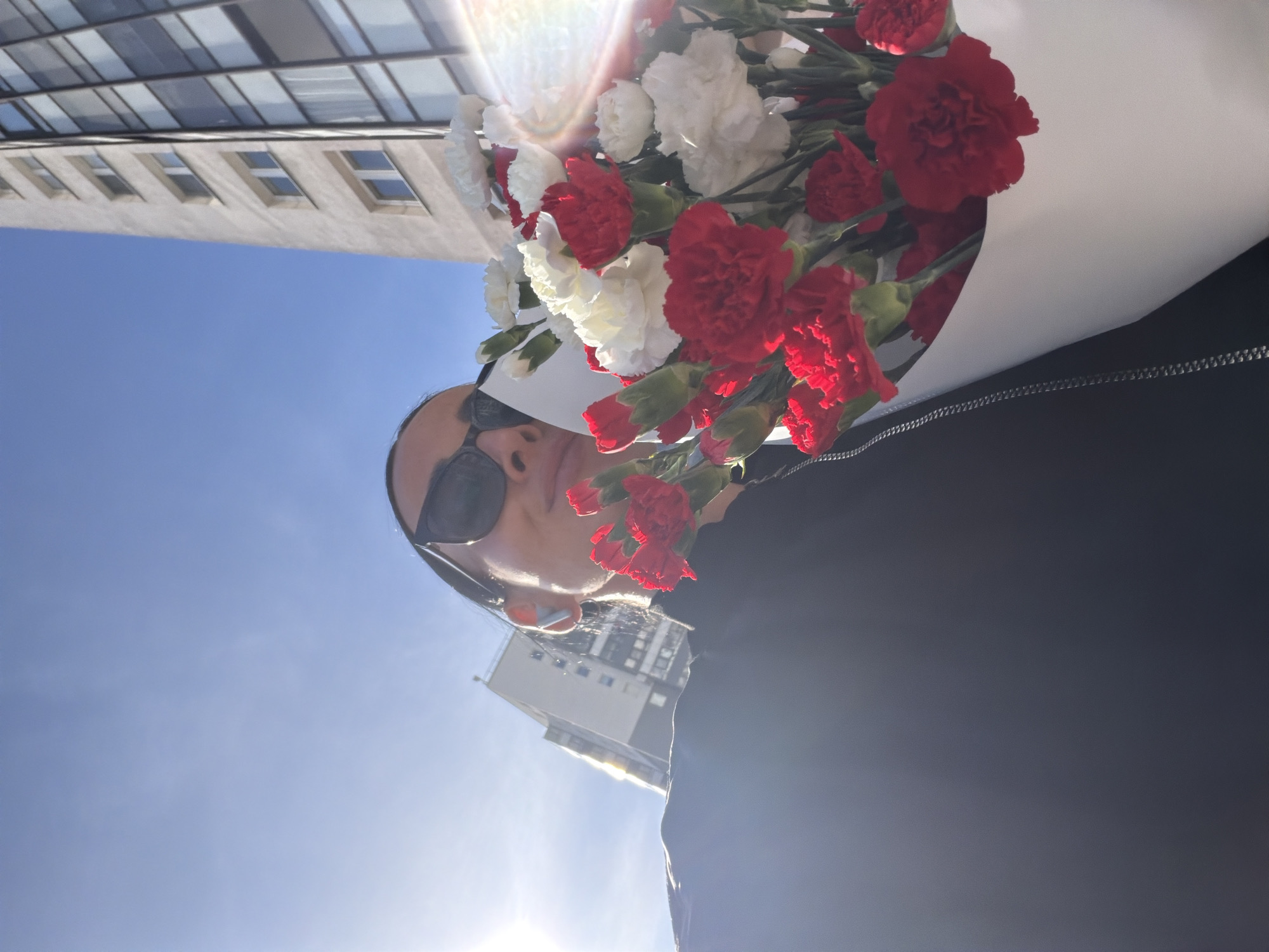 A person wearing sunglasses holds a bouquet of red and white flowers in front of them, partially obscuring their face. The background shows a bright blue sky and a tall building with multiple windows. The location is Saint Petersburg, Russia, as inferred from the architectural style of the building.