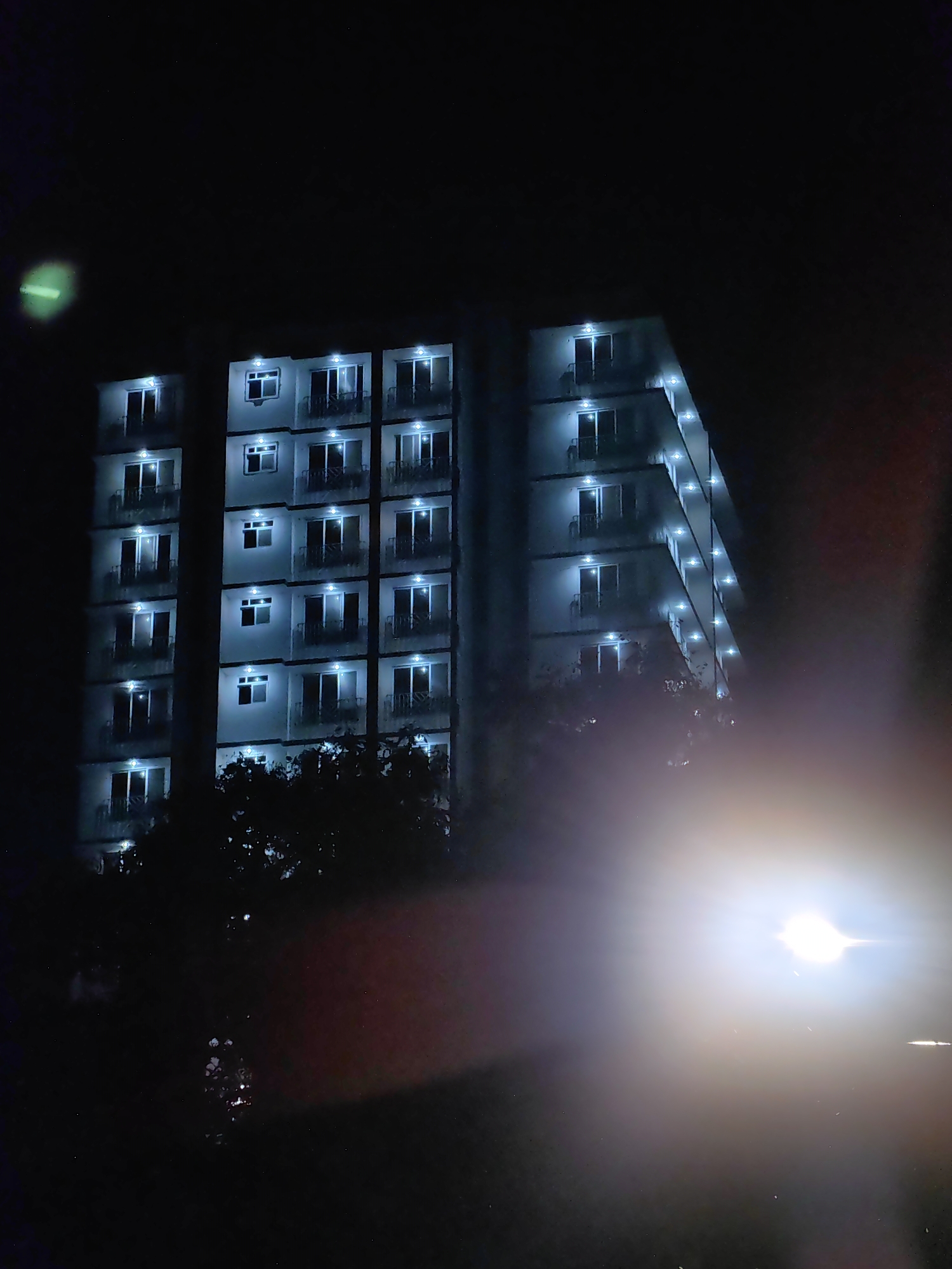 The image shows a modern, multi-story apartment building at night in Mumbai, India. The building is brightly lit from within, with rows of windows and balconies illuminated. Trees and foliage are visible at the base of the building. The foreground is dark and out of focus, with a bright light source, possibly a street lamp or car headlight, creating a lens flare effect. The overall impression is one of urban nighttime activity.