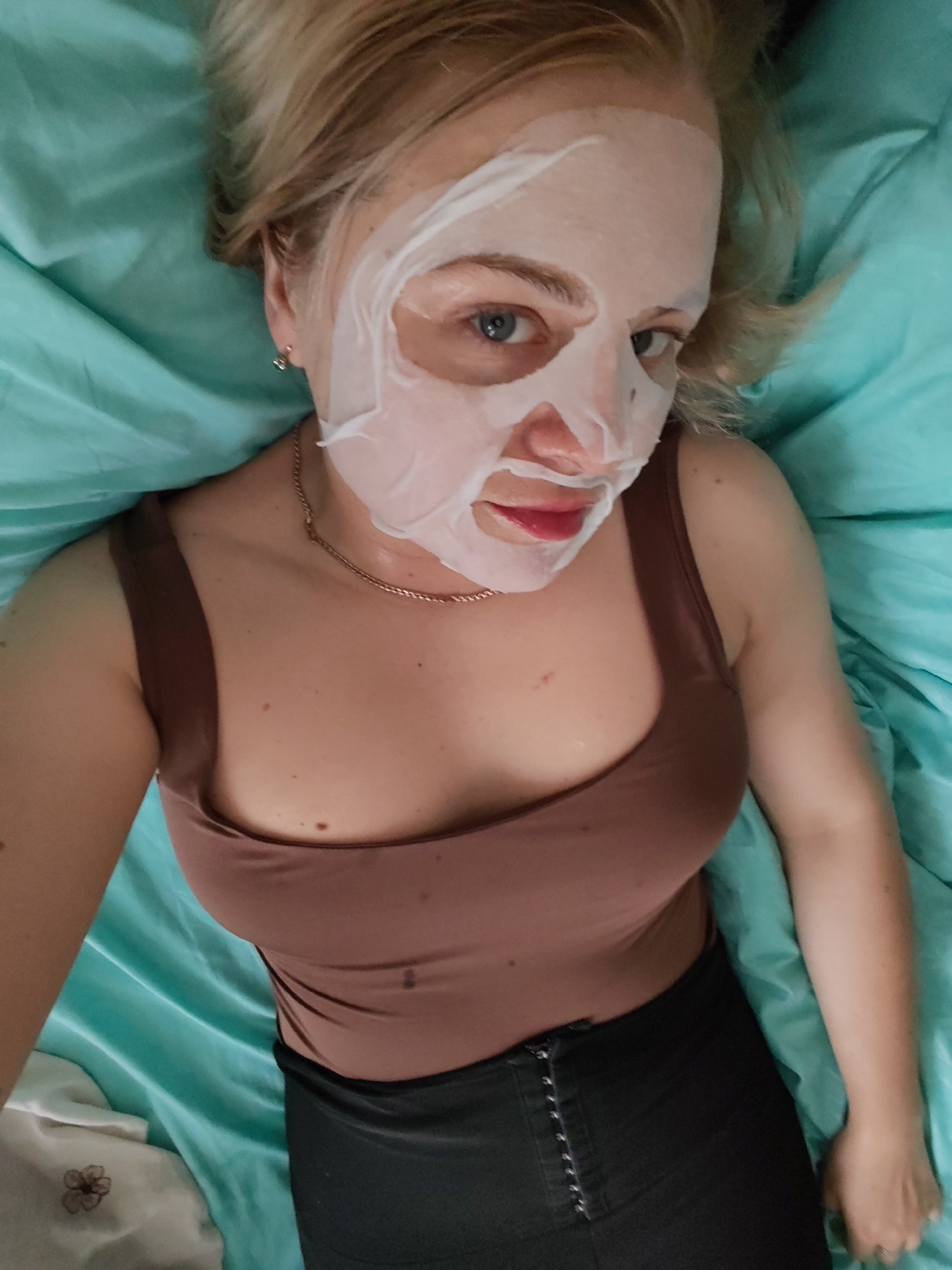 A woman with blonde hair is lying on a bed covered with turquoise sheets. She is wearing a brown tank top and black pants. A white sheet mask covers her face, leaving her eyes and lips visible. A thin gold chain necklace is around her neck. The scene is an indoor setting, likely a bedroom. There is no information available in the image to determine the location within the city of Izhevsk, Russia.