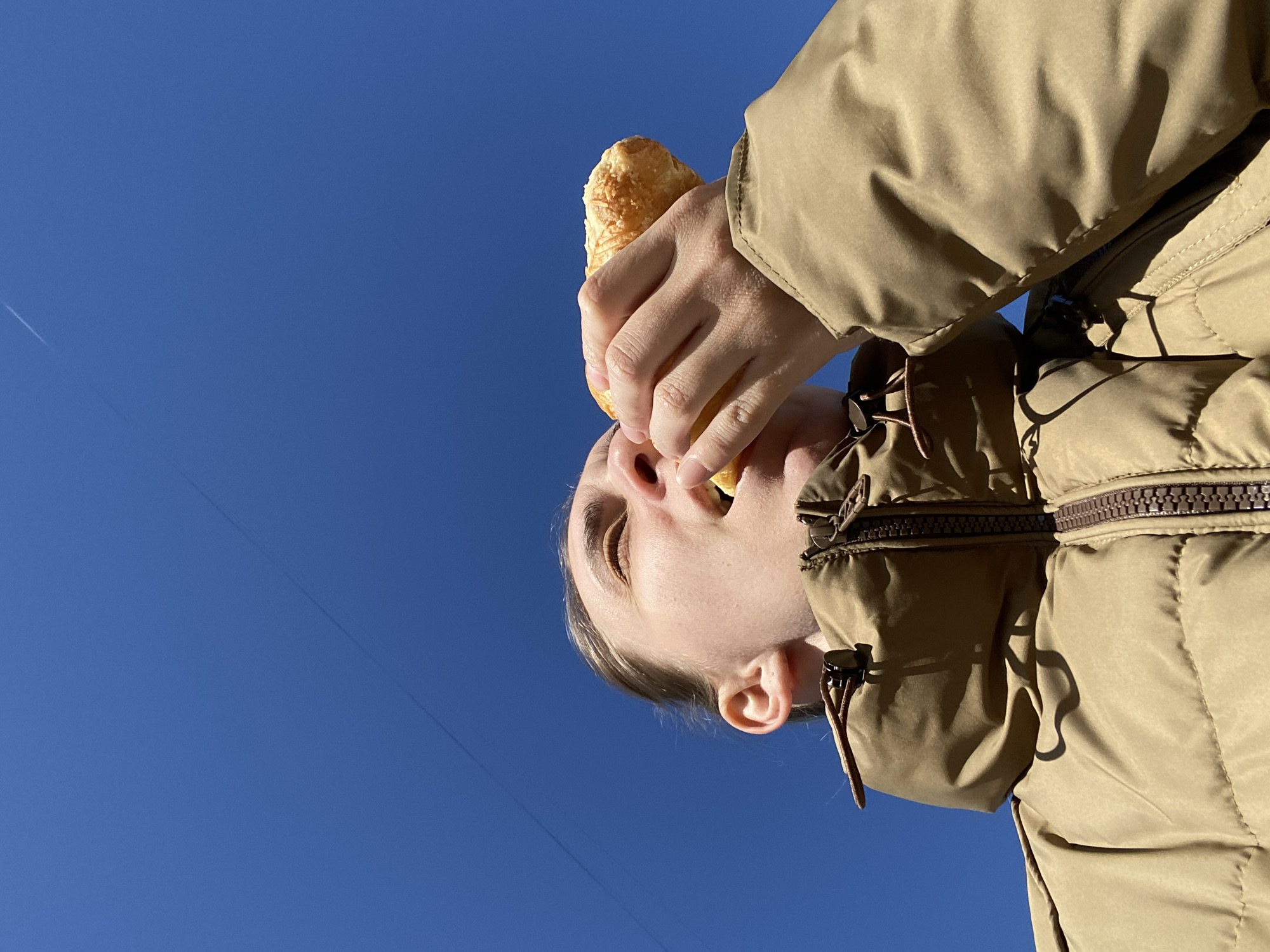 A low-angle, rotated view of a person eating a pastry against a cloudless, blue sky. The person's face is visible, and they are holding the pastry with their hand near their mouth. The person wears a light brown, quilted jacket. A faint contrail is visible in the blue sky. There is no contextual information suggesting the photo was taken in Chaikovsky, Russia.