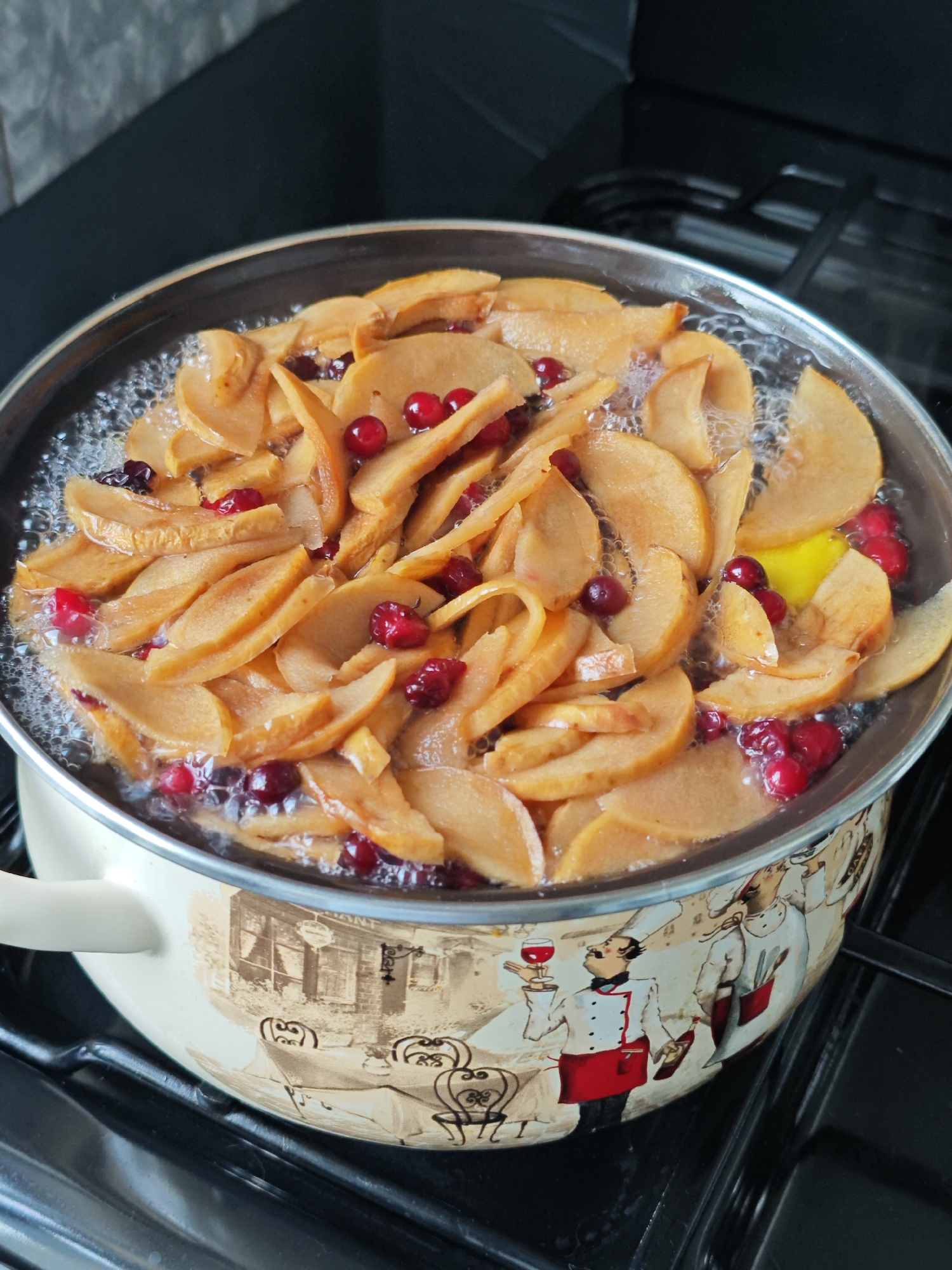 A medium shot captures a pot of fruit simmering on a gas stovetop. The pot, cream-colored with a decorative band depicting chefs and a café scene, is filled with sliced apples and whole cranberries in a bubbling liquid. A slice of lemon is also visible among the fruit. The cooking process appears to be in full swing, with visible steam and bubbles on the surface of the liquid. The background consists of a dark stovetop and a dark countertop, with a hint of a grey tiled backsplash. The lighting is bright and even, suggesting indoor illumination. No specific time of day or weather conditions can be inferred. The visible text on the pot is part of the decorative design and is not legible. The overall impression is that of a home kitchen environment where a comforting dessert or beverage is being prepared. The scene is static, focusing on the culinary activity.