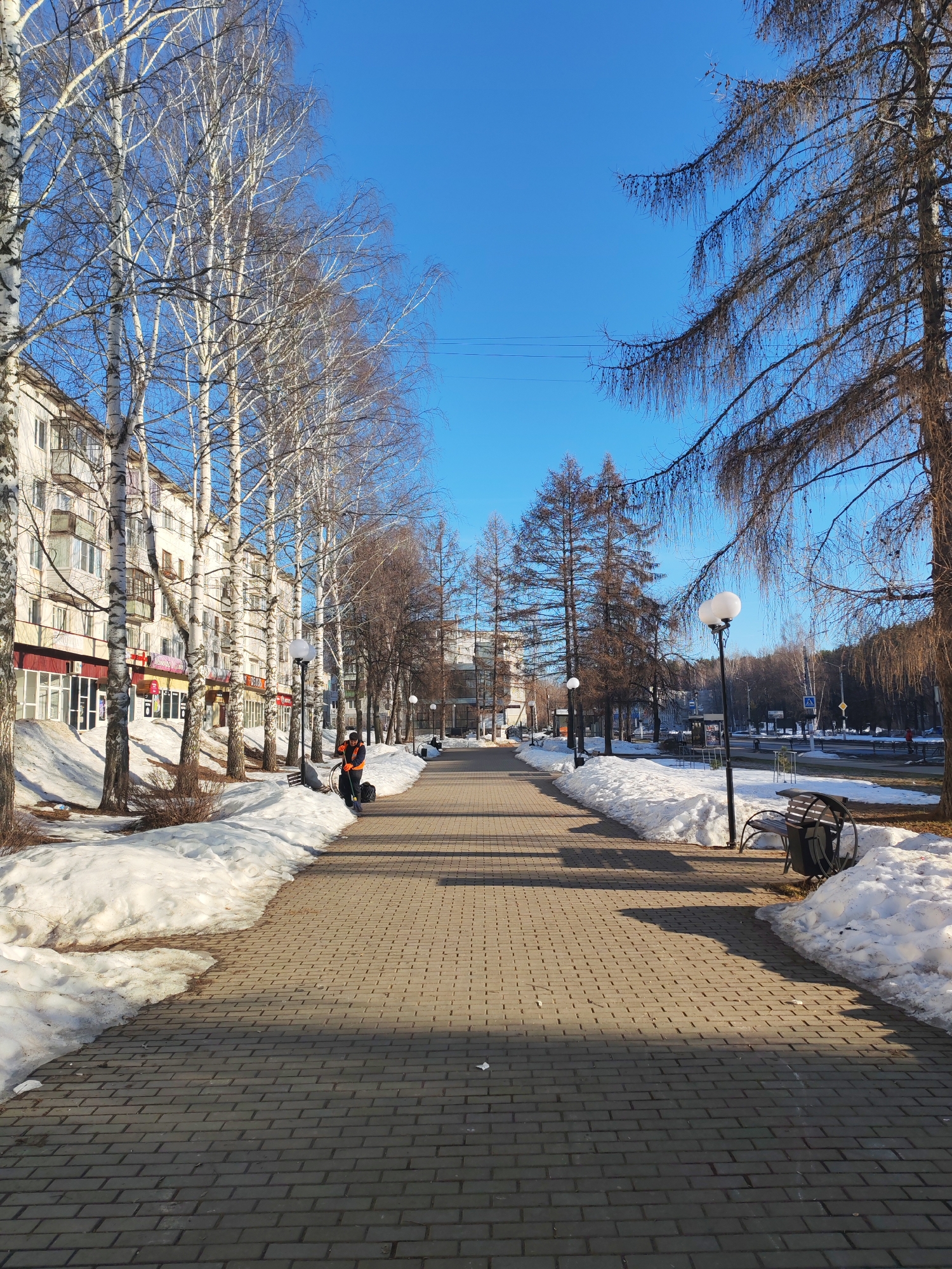 The image depicts a winter scene in Chaikovsky, Russia. A paved walkway lined with trees and benches stretches into the distance, with buildings visible on the left side. Patches of snow remain on either side of the walkway. A person dressed in an orange jacket is sweeping the walkway. Overhead, the sky is clear and blue. Streetlights are spaced along the walkway.
