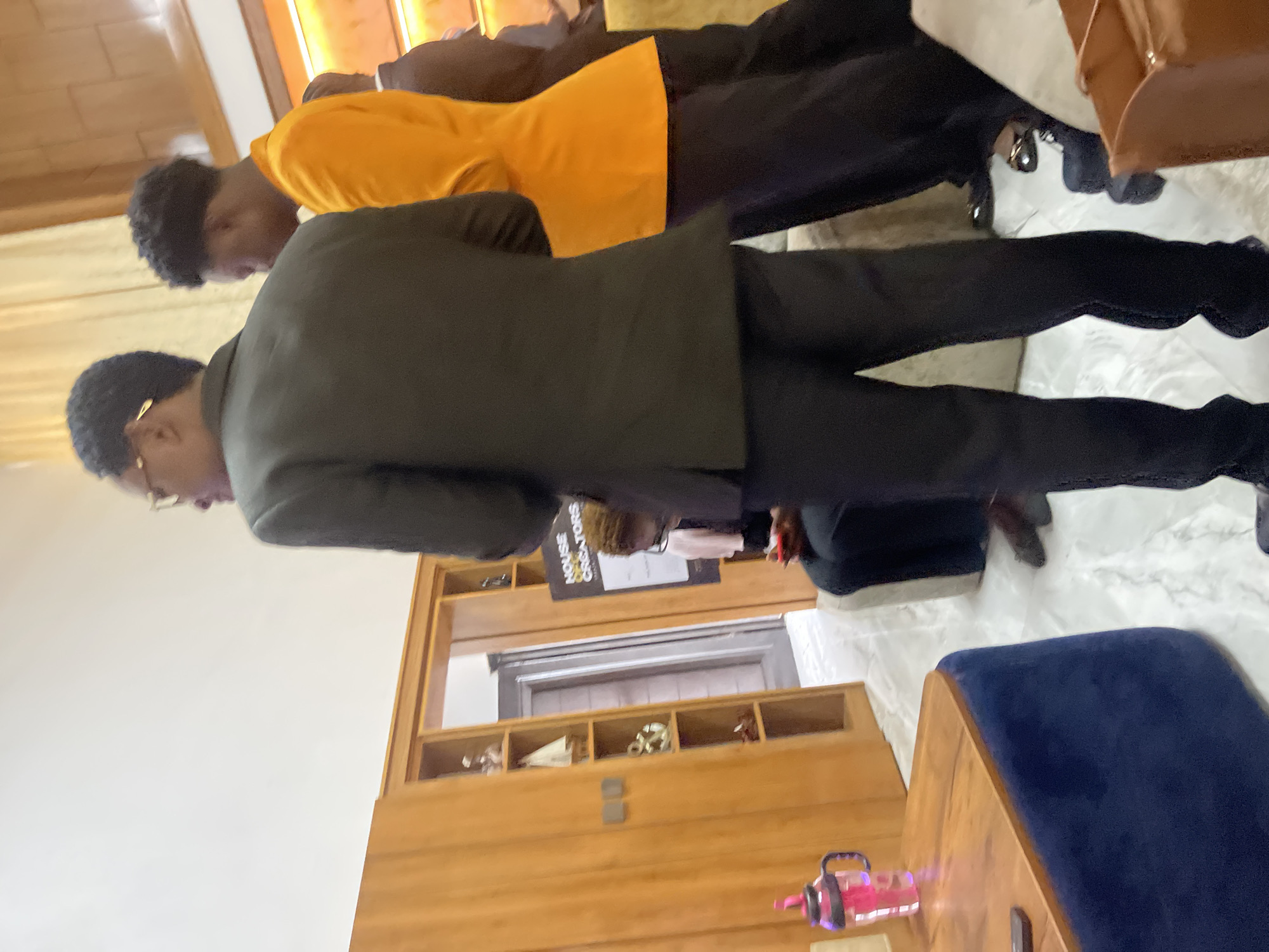 The image depicts an indoor scene, possibly in Gura Top, Nigeria, with a group of individuals. In the foreground, a person is partially visible from the back, wearing a dark suit. To their right, another individual is seen from the side, dressed in a bright orange jacket and dark pants. Further back, a person in a dark suit is standing, and another individual in dark clothing is seated, looking at a phone.
The environment includes a wooden cabinet with shelves displaying decorative items, a closed wooden door, and a sign that reads "HOUSE CREATORS". A wooden table with a blue upholstered base is also present, along with a pink water bottle. The floor appears to be marble. The lighting suggests an indoor, possibly modern, setting. The overall impression is of a gathering or event.