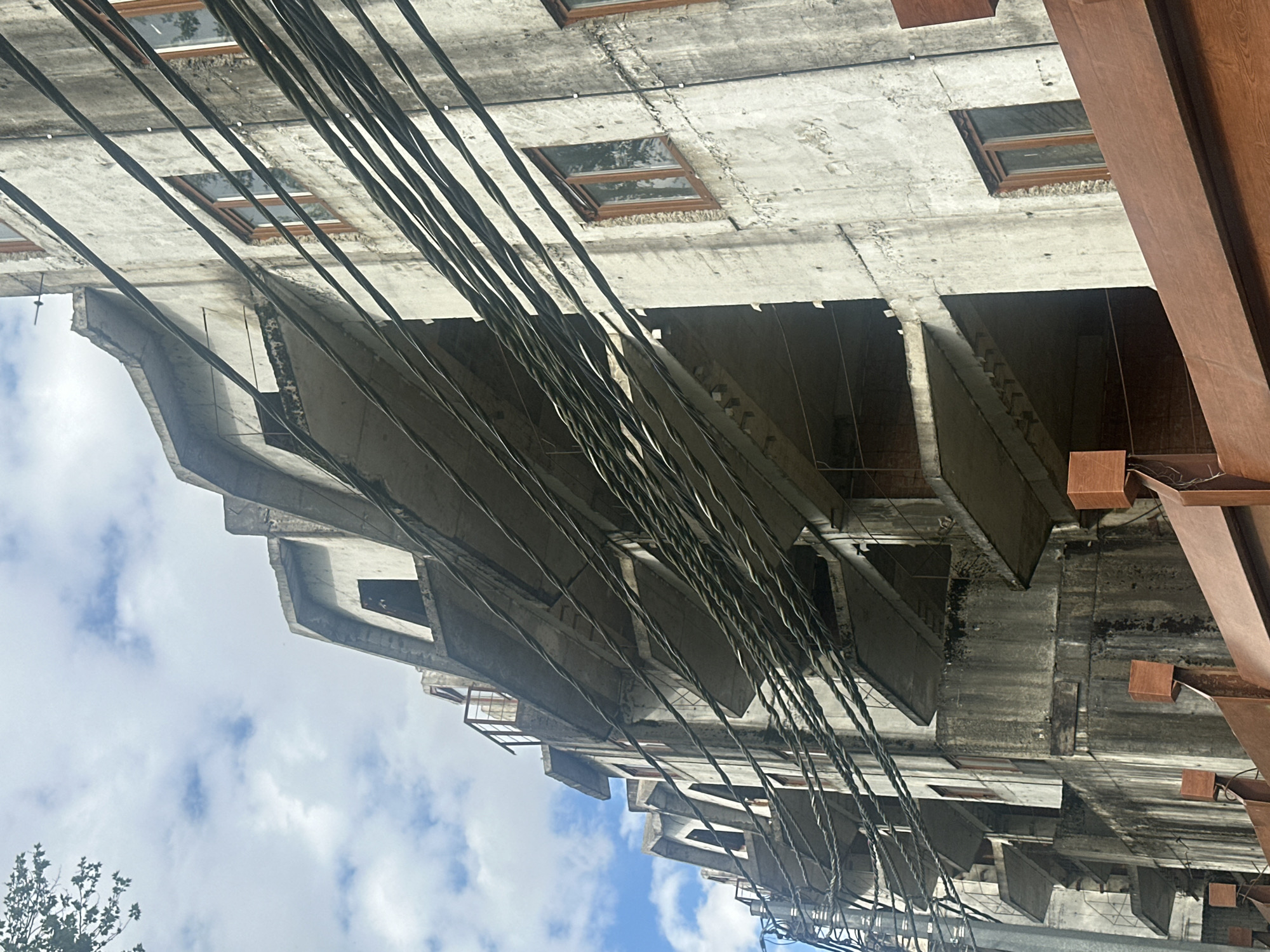 The image shows an upward view of a multi-story concrete building in Krasnaya Polyana, Russia. The building exhibits exposed concrete features and a series of cantilevered platforms. Numerous power lines and cables run diagonally across the foreground, partially obscuring the view of the structure. Small, rectangular windows are visible along the top portion of the building. The sky, with scattered clouds, is visible in the lower-left corner. A section of rusted steel or iron framing is visible on the right.