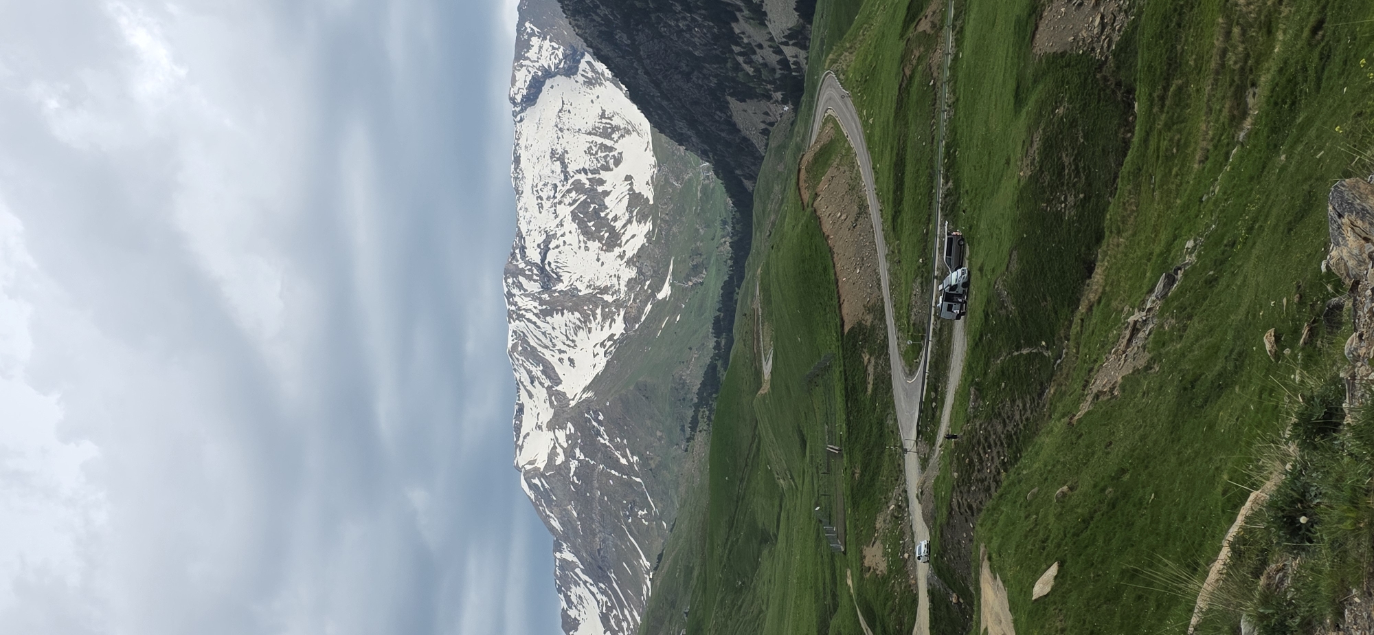 The image captures a scenic view of a mountain pass in Benasque, Spain. A winding road ascends through lush green hillsides, with vehicles parked along the roadside. A snow-capped mountain peak rises in the background, contrasting with the verdant landscape. The sky overhead is overcast with gray clouds.