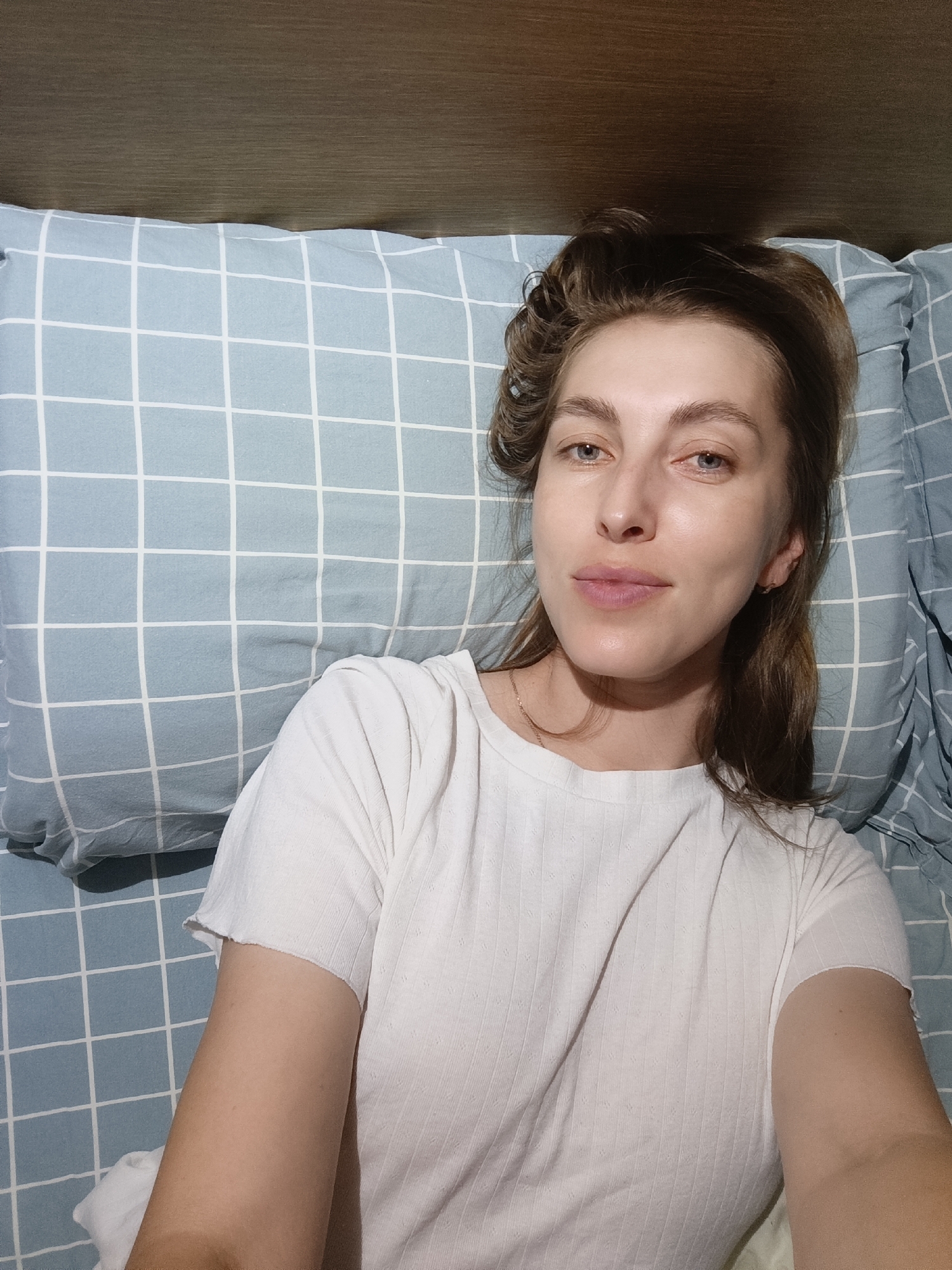A woman with brown hair and blue eyes is lying in bed. She is wearing a white t-shirt and is on a blue and white checkered pillow. The background is a wooden headboard. The image appears to be taken indoors in a bedroom. The location is in the city of Chaikovsky, Russia.