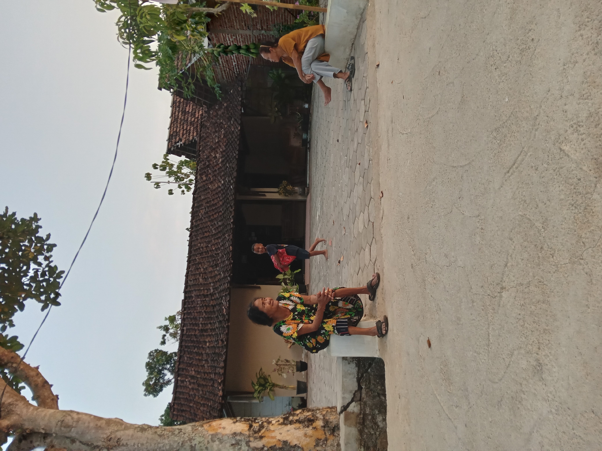 In Kedunggudel, Indonesia, an outdoor scene depicts three individuals in front of a traditional Indonesian house with a tiled roof. On the left, a woman with dark hair, wearing a vibrant floral dress and sandals, sits on a concrete ledge. Her hands are clasped in her lap, and she looks to her right. To her right and further back, a child in a dark shirt and shorts is walking away from the camera, carrying a red object. On the far right, another person, wearing an orange t-shirt and grey pants, sits on a similar concrete ledge, with their legs extended. This individual is looking towards the left. The ground in front of the house is paved with hexagonal stones. A large tree trunk is visible on the far left, and lush green foliage is present on the right side of the image. The sky is clear and pale.