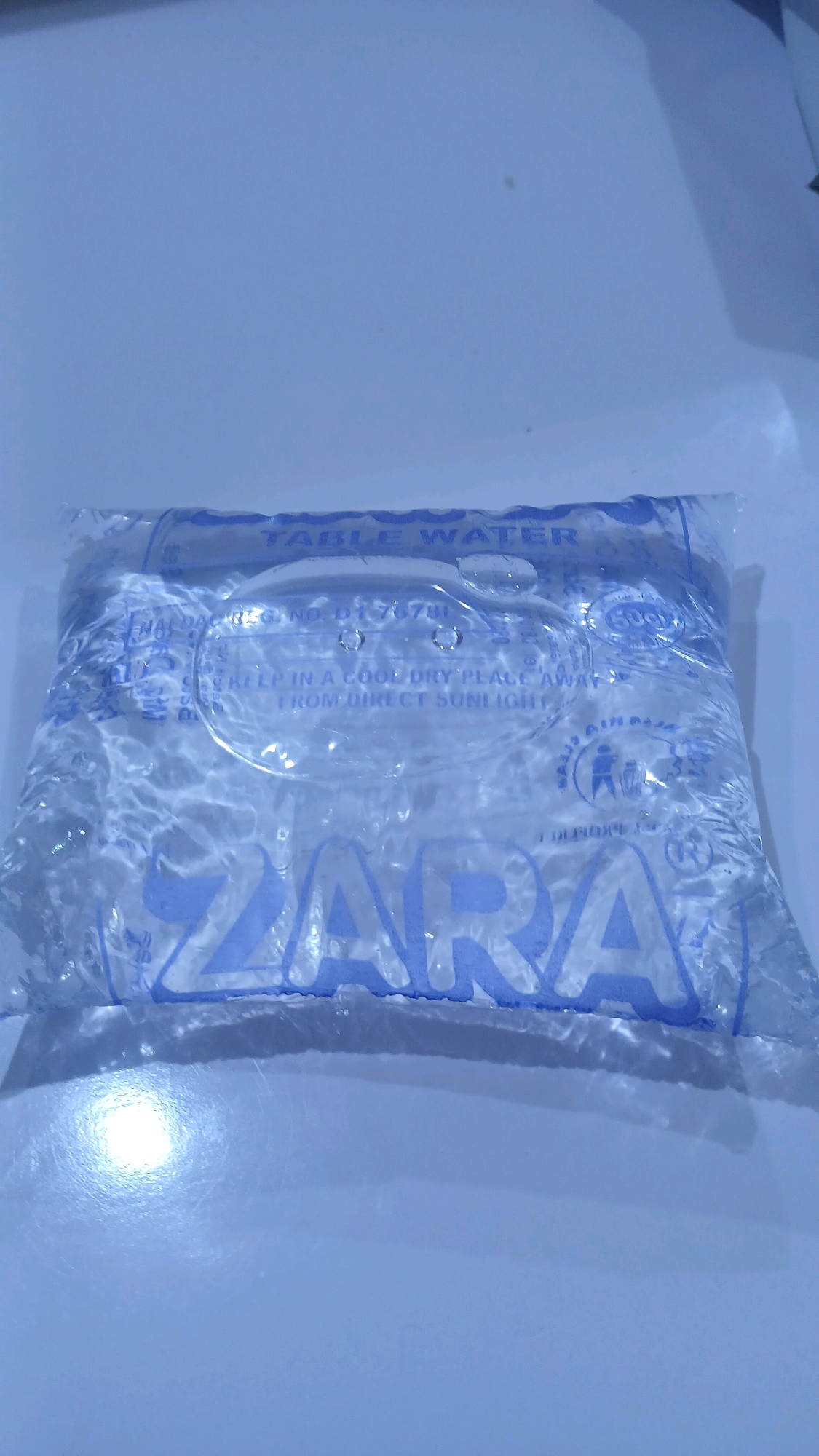 The image shows a bag of table water on a white surface. The bag is clear plastic and filled with water, with ripples distorting the view of the label. The label is blue and white, and some text is visible, including "TABLE WATER" and "ZARA". There is also a registration number and instructions to "KEEP IN A COOL DRY PLACE AWAY FROM DIRECT SUNLIGHT". The background is a plain white surface, possibly a table. The lighting is bright and creates a glare on the water. The location is identified as Gura Top, Nigeria.