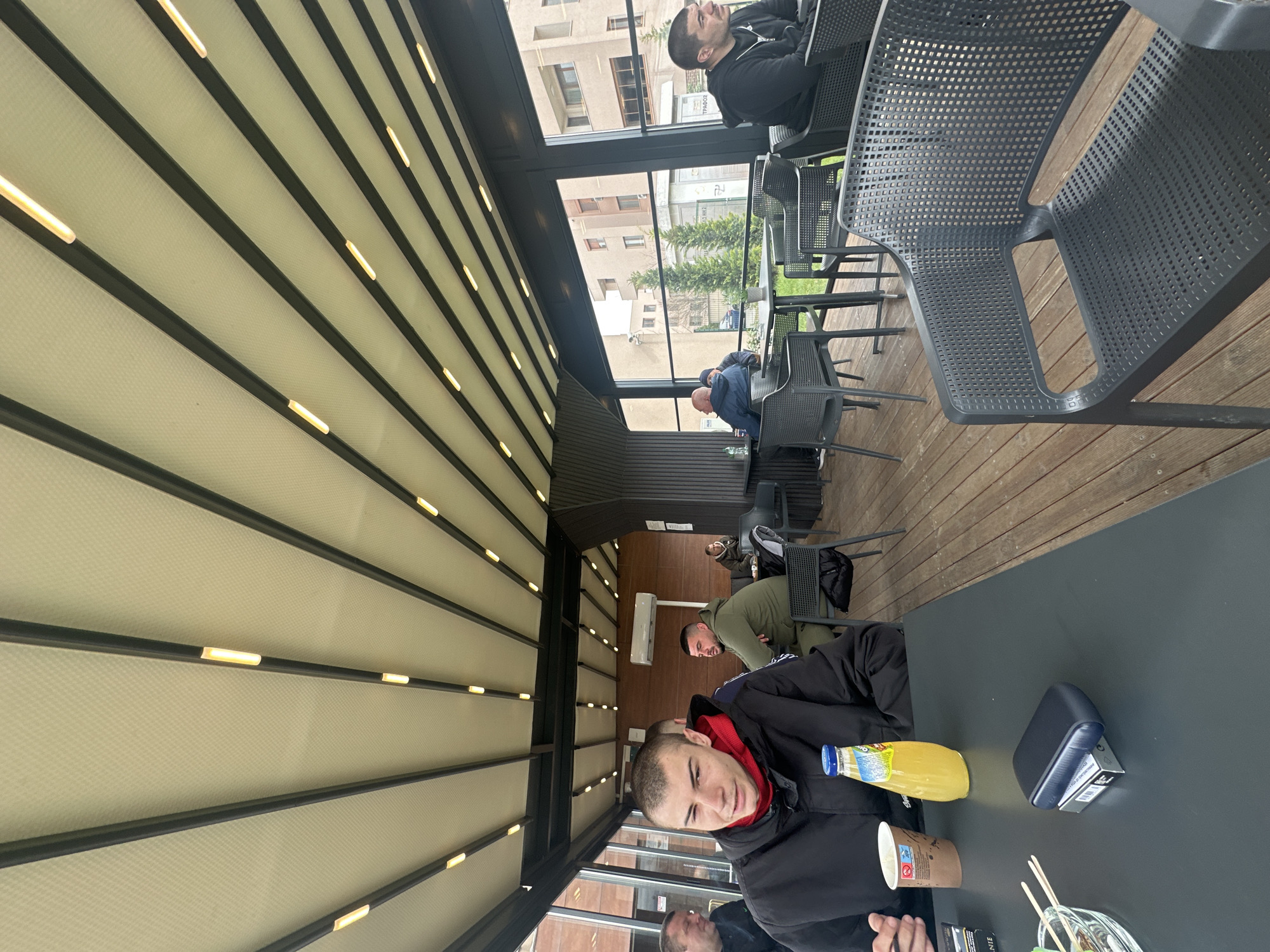The image depicts a group of men in an outdoor cafe setting. A man sits at a dark-colored table where a juice bottle, a disposable cup, a pack of cigarettes, and a glass dish with what appears to be food or a beverage are present. He is wearing a black jacket with a red hood.
Other men are seated at tables further away, some appearing to be deep in conversation. The cafe has a retractable roof made of a white material with black support beams with lighting elements. Outside the cafe is a view of buildings in a city.
Based on the architectural style and visible signs in other photographs taken at the same location, it can be assumed that the location is in the city of Plovdiv, Bulgaria. The cafe has dark grey chairs with a perforated design. The flooring appears to be made of wooden planks.