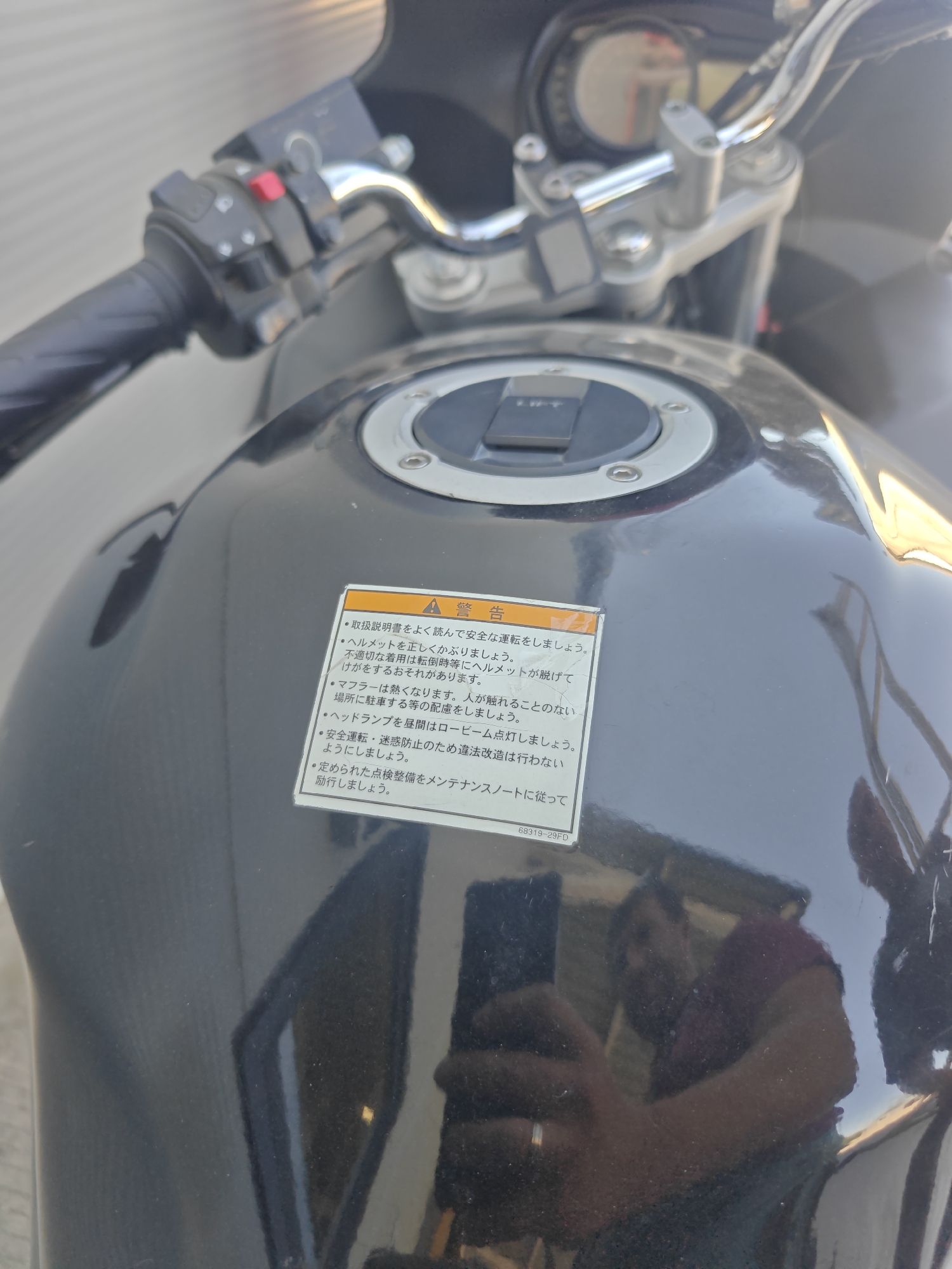 The image shows a close-up of a motorcycle's fuel tank and handlebars. A warning sticker is prominently displayed on the fuel tank, written in Japanese. The sticker contains several points of advice related to safe motorcycle operation, including wearing a helmet, being aware of the hot exhaust, ensuring the headlight functions correctly, and adhering to maintenance schedules. The background appears to be an outdoor setting with a slightly blurred effect, suggesting it was taken in an urban environment, possibly in Krasnodar, Russia, though no definitive landmarks are visible. The lighting indicates it is daytime. A reflection of a person holding a phone is visible on the motorcycle's surface. The motorcycle is black and appears to be in good condition.