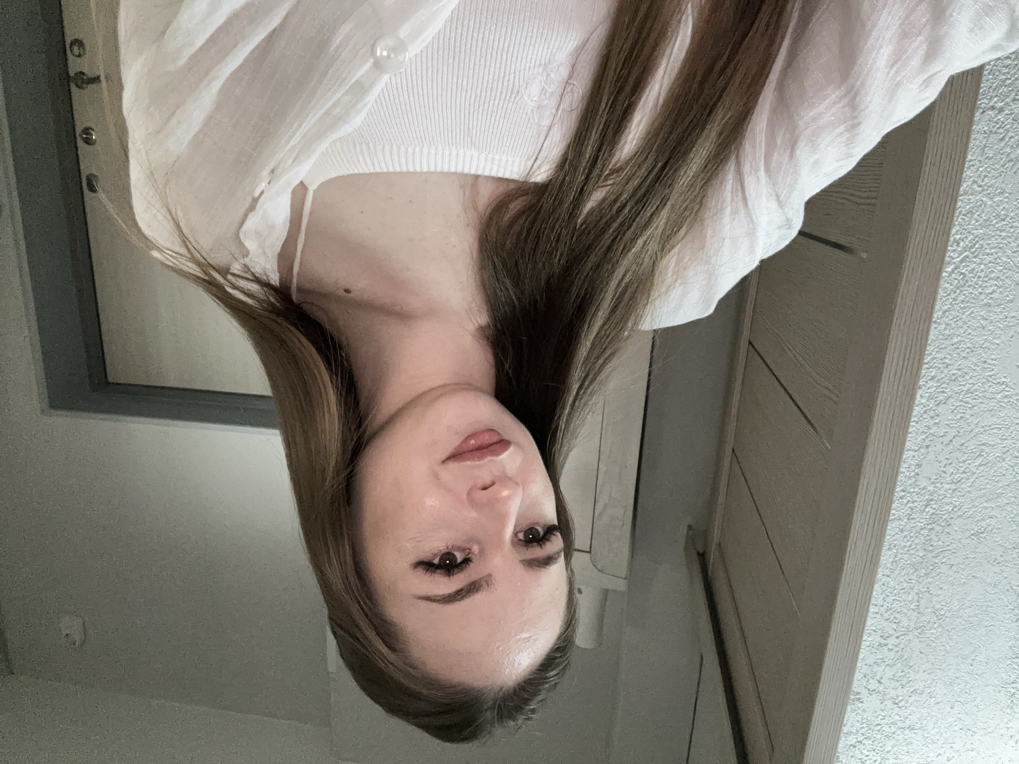 A young Caucasian woman with long, straight brown hair and fair skin is shown from the chest up, looking to her right. She is wearing a white ribbed tank top and an unbuttoned white linen shirt. Her makeup includes foundation, blush, mascara, and lip gloss. The background appears to be an indoor setting with textured white walls and a wooden door. The lighting is dim, suggesting an indoor environment at dusk or night. The image is a selfie taken from a low angle.