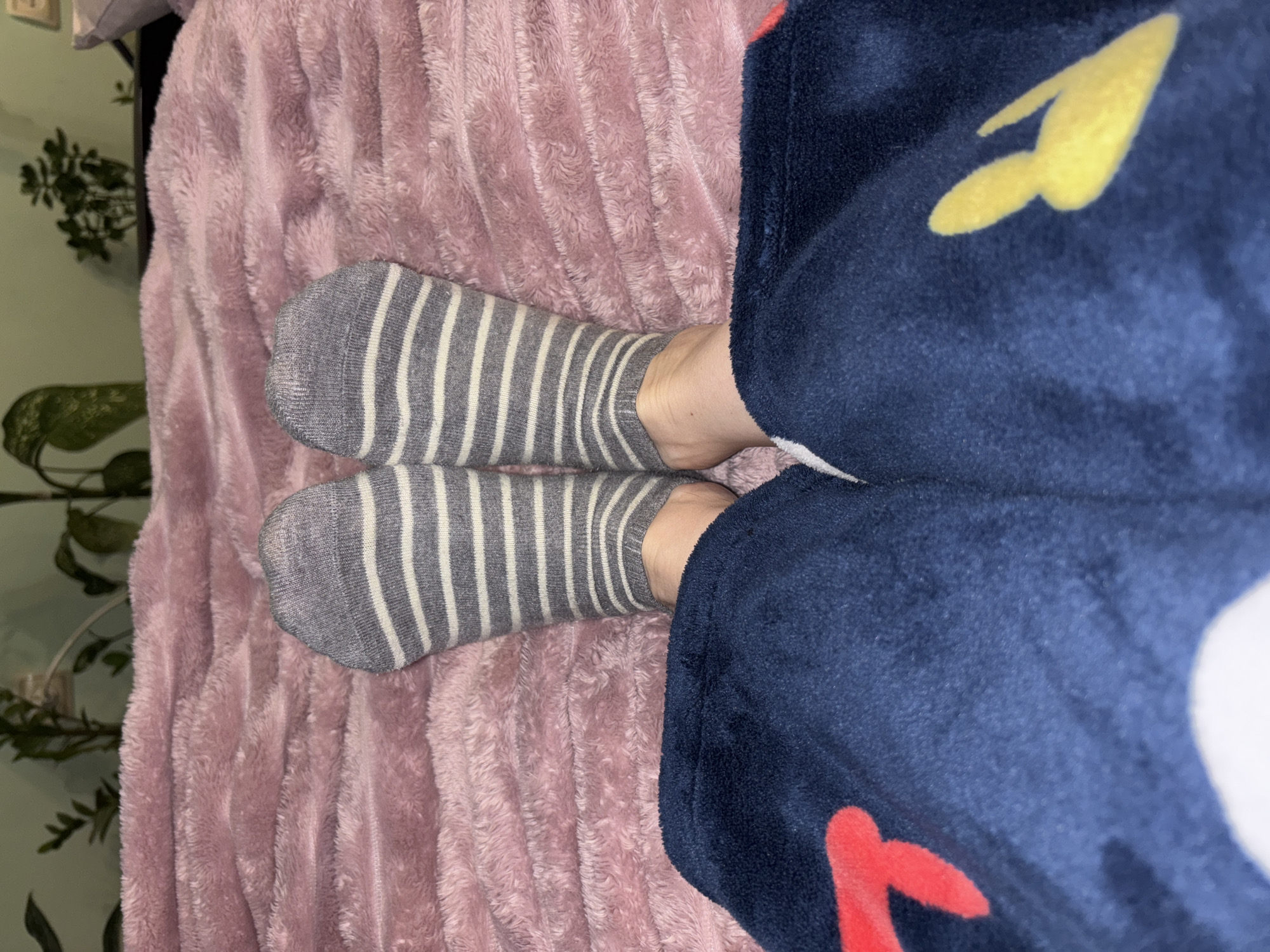 The image shows a person's legs and feet on a pink blanket. The person is wearing gray socks with white stripes and blue pajamas with a colorful pattern, possibly lightning bolts and flames. The scene appears to be indoors, potentially in a bedroom. The location is Unknown, Russia.