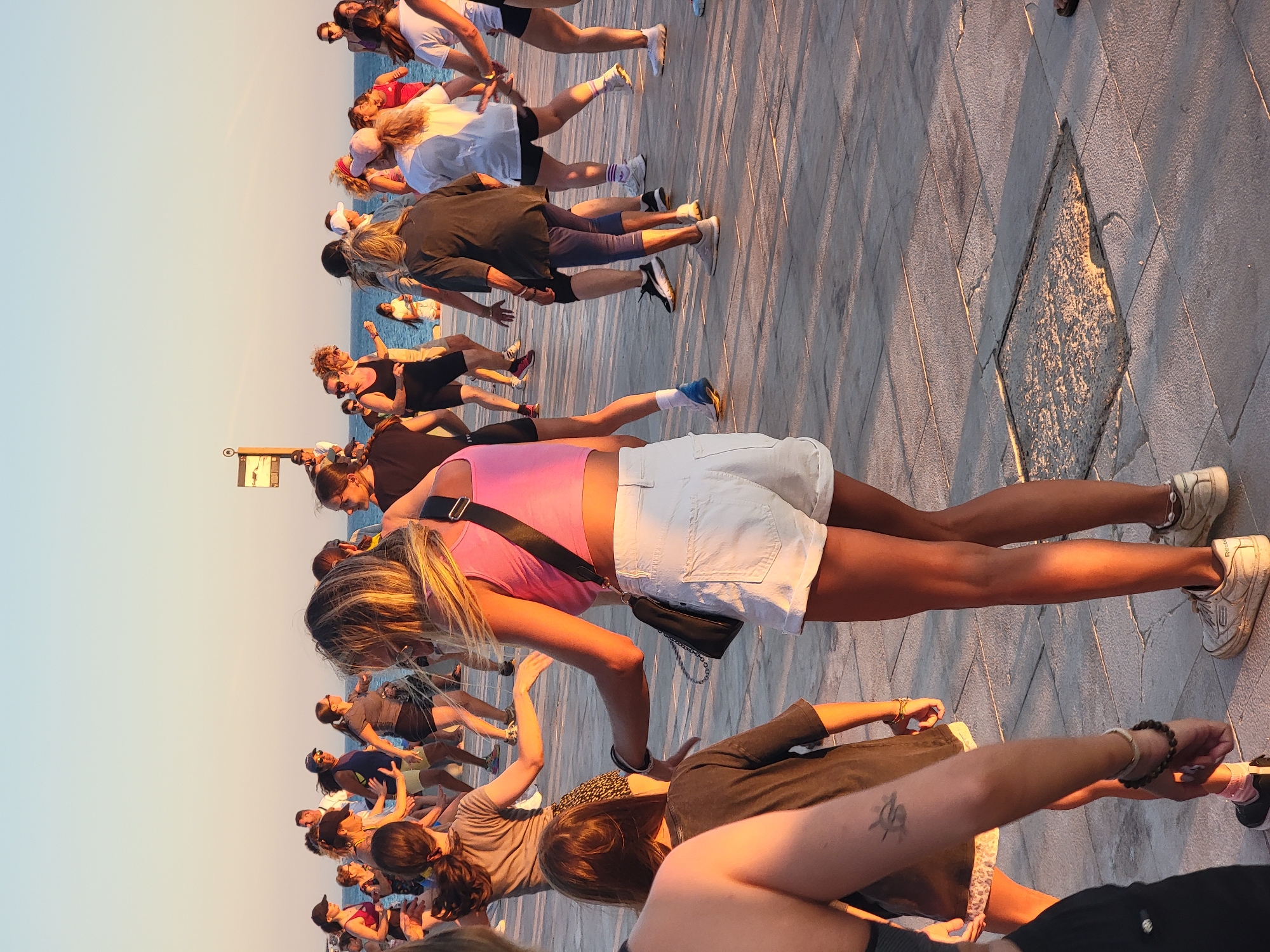 The image captures a group of people engaged in what appears to be an outdoor dance or fitness class on a paved promenade in Quarteira, Portugal. The scene is illuminated by the warm, golden light of late afternoon or early evening, casting long shadows across the stone-tiled ground. The ocean is visible in the background under a clear, pale blue sky. The individuals, a mix of adults and at least one child, are dressed in casual, athletic attire. They are in various poses, suggesting movement and participation in a synchronized activity. One woman in the foreground, wearing a pink crop top and white denim shorts, is turned away from the camera, her arms extended. To her left, a child in a dark t-shirt looks towards the activity. Other participants are scattered throughout the frame, their postures indicating engagement in a dance or exercise routine. A signpost is visible in the distance. The overall atmosphere is one of energetic recreation in a public, seaside setting.