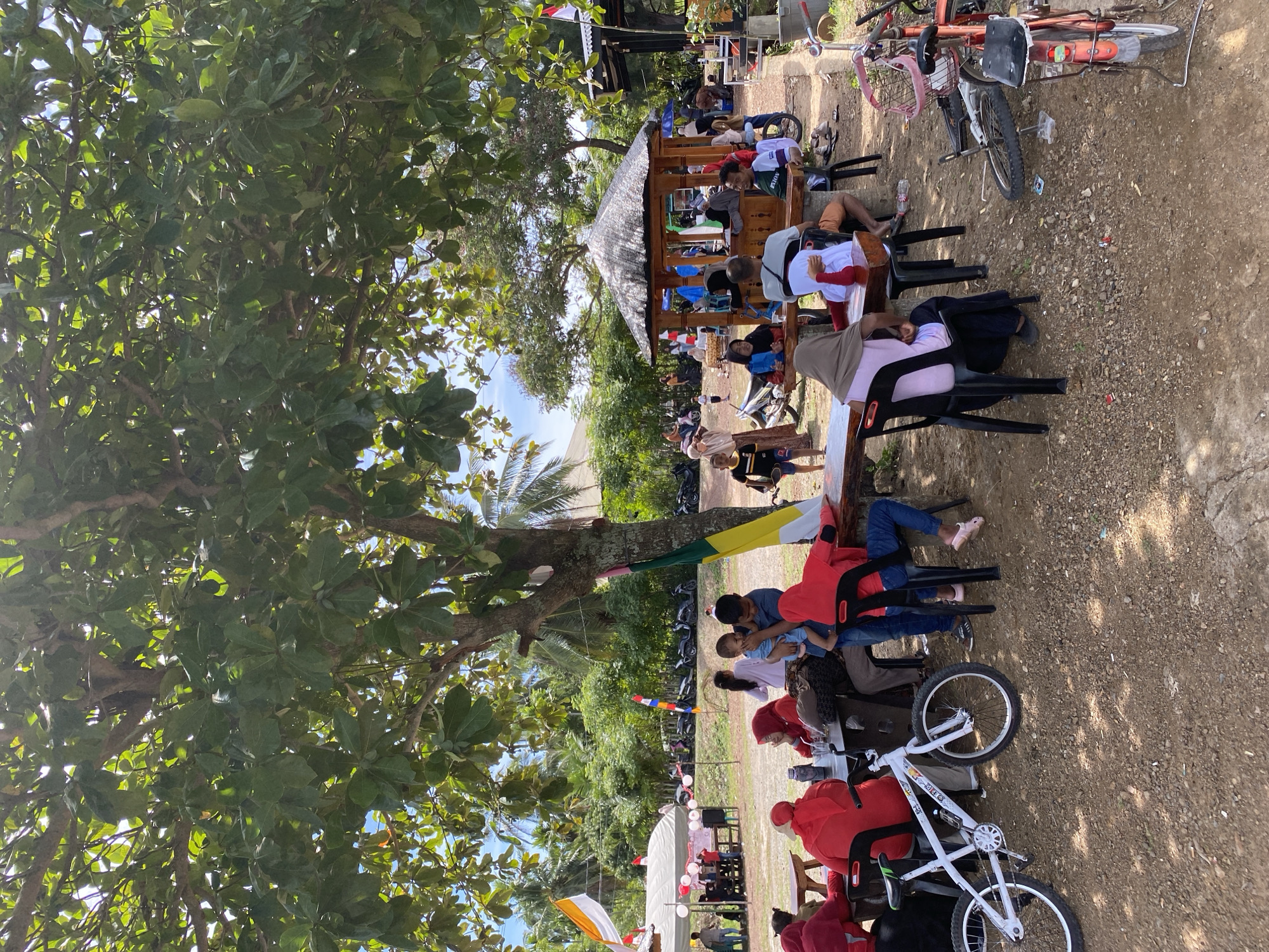 In Aneuk Paya, Indonesia, this outdoor scene depicts a gathering of people seated at tables under the shade of a large tree. The ground is covered with dirt and small stones. In the foreground, a white BMX bicycle is parked, and a red bicycle with a basket is visible on the right side of the image. Several people are seated at wooden tables, some wearing red clothing and headscarves, others in casual attire. A small gazebo with a thatched roof is in the mid-ground, with more people gathered around it. In the background, there is lush greenery with palm trees, indicating a tropical environment. Several motorcycles are parked in a line. Flags are also visible, including one with orange and white stripes and another with green, yellow, and red. The sky is bright blue with some clouds, suggesting a sunny day. The overall impression is of a relaxed community event or a casual outdoor dining area.