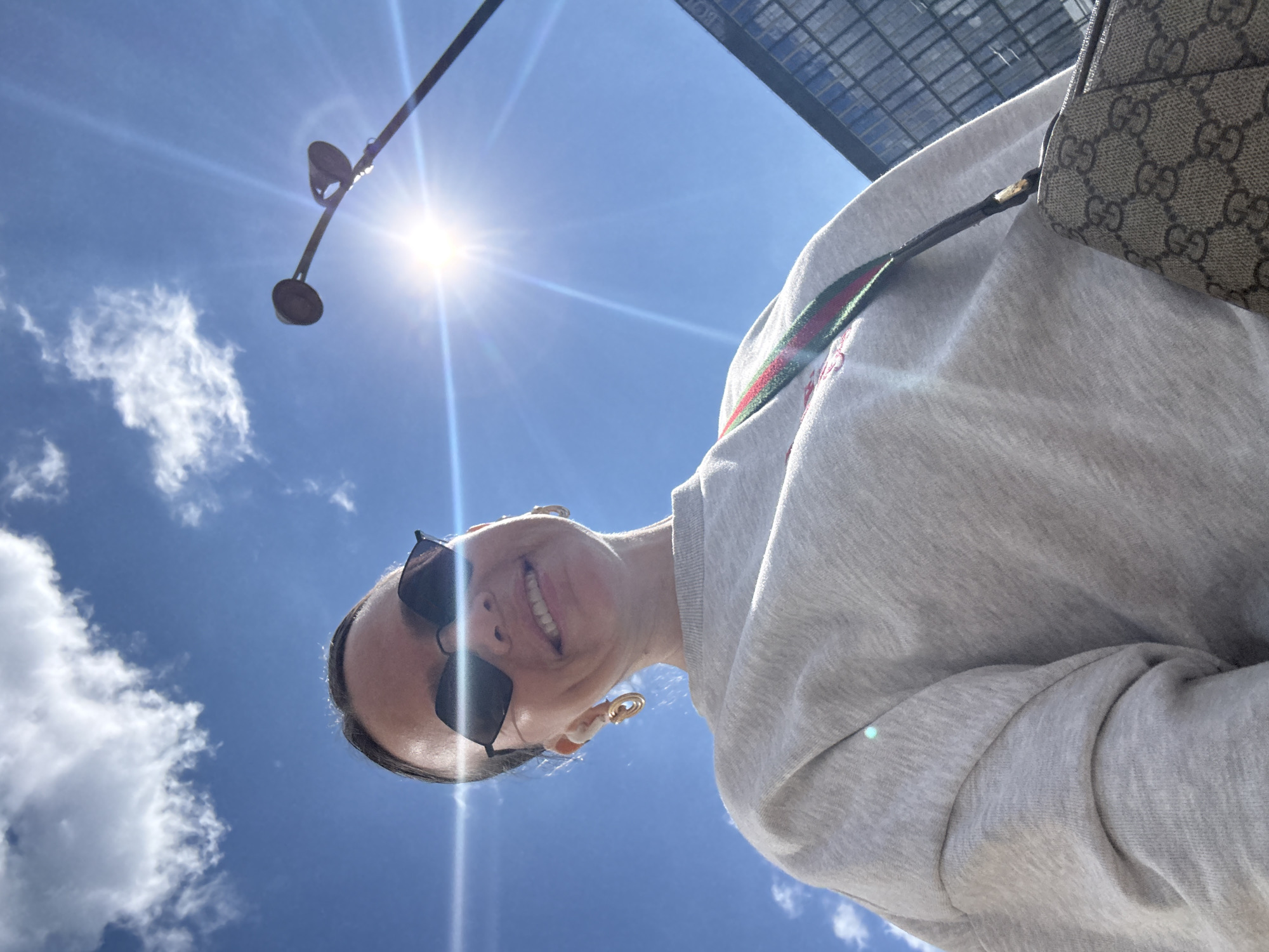 A woman wearing sunglasses and a light-colored sweatshirt takes a selfie against a bright blue sky with scattered clouds. She is also wearing gold hoop earrings and a purse with a distinctive pattern. The sun is shining brightly, creating lens flare. In the background, a modern building with a glass facade is visible. The scene suggests an outdoor location in a city.
