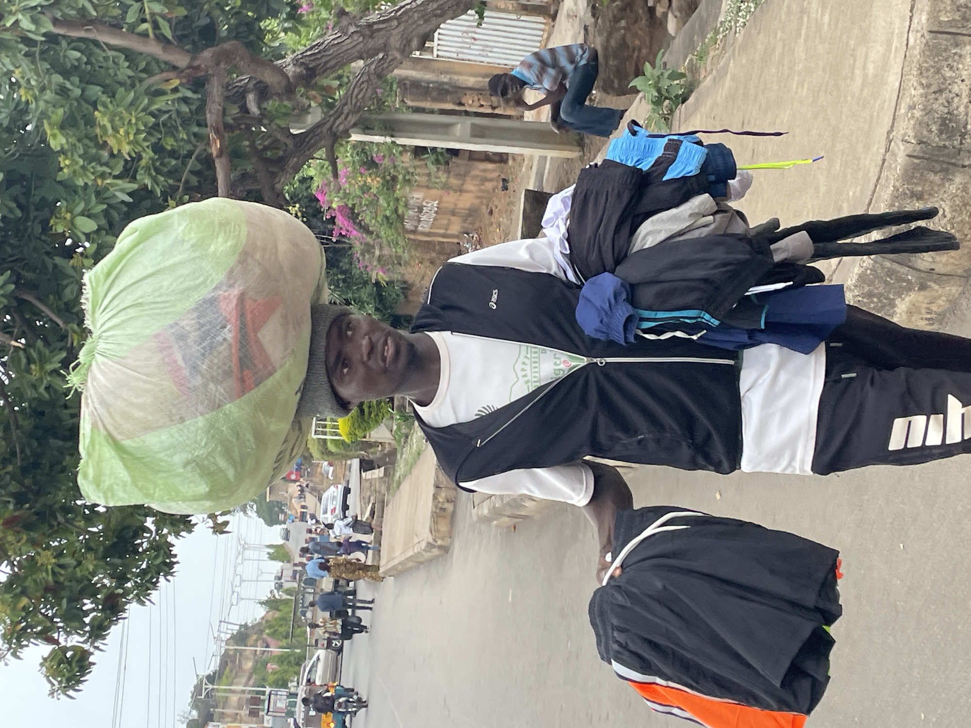 The image displays a daytime street scene, likely in an urban or semi-urban area of Jos, Nigeria. The central figure is a dark-skinned man, appearing to be a street vendor, standing on a paved surface and looking directly at the camera. He wears a dark beanie, a black athletic jacket with a white "ASICS" logo on the left chest, a white t-shirt with faint green text (including "HAPPEN" and "GER," possibly part of "NIGERIA"), and black shorts bearing a white "mhh" logo on the right leg.
He is carrying a large, light green mesh bag, filled with various items (likely clothing, with visible red, orange, and white patterns), balanced steadily on his head. Additionally, he has a significant bundle of clothes, including black, blue, white, and grey garments, draped over his left shoulder and arm. Some hangers are discernible within the bundle.
In the background, the street extends, populated by several pedestrians, a motorcycle, and cars, suggesting a busy thoroughfare. To the man's right, another individual wearing a striped shirt and dark pants is visible crouching or sitting on a low concrete wall. Lush green trees and foliage frame the left and upper portions of the image. The sky appears overcast or hazy, providing soft, diffused daylight. The man's expression is neutral and stoic. The scene captures a snapshot of daily life and informal commerce.