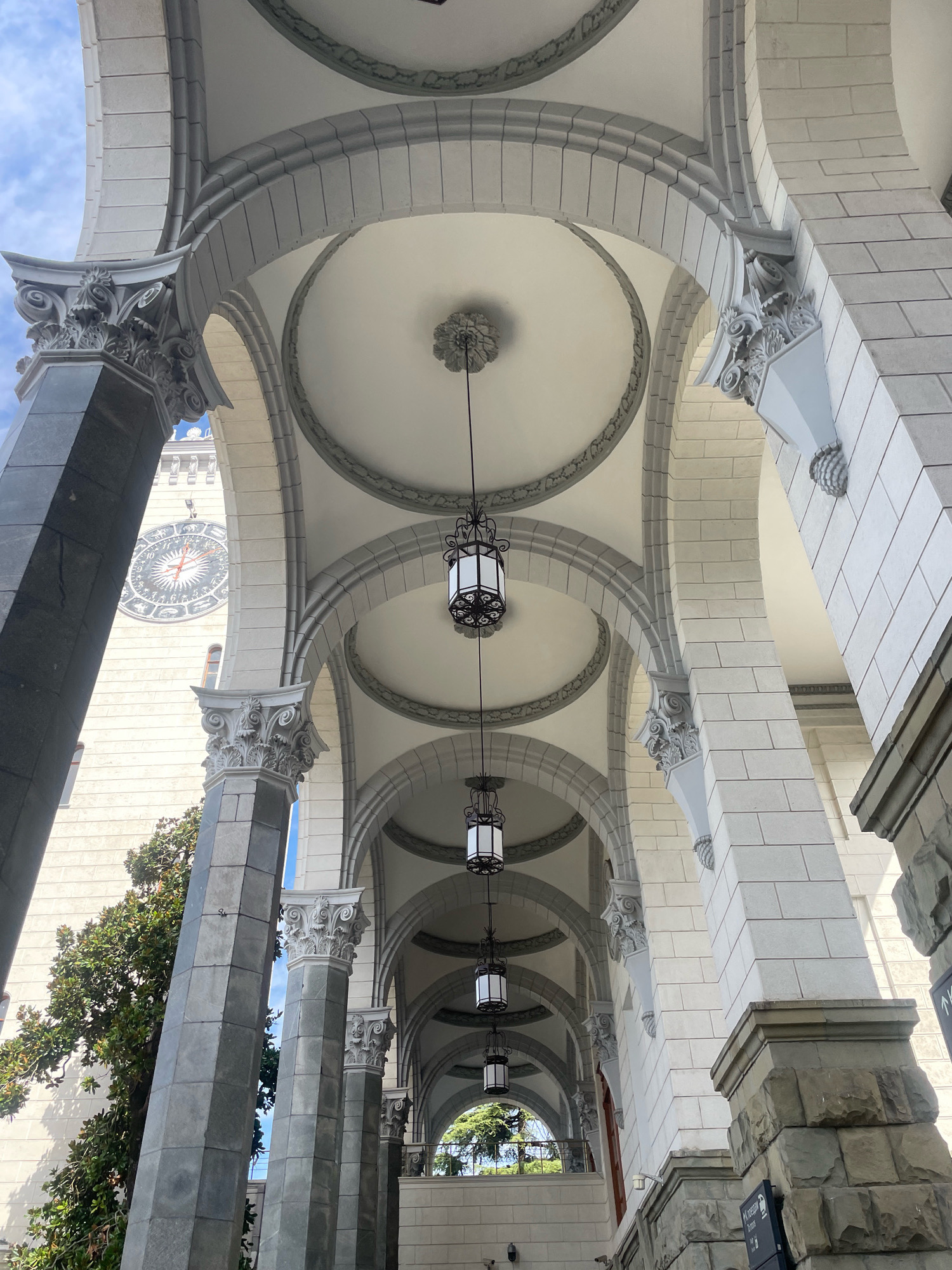 An exterior, low-angle, eye-level shot depicts a grand arched walkway of a building, possibly a historical landmark, with ornate architecture. The walkway is characterized by a series of symmetrical arches, supported by thick, stone pillars, and a decorative vaulted ceiling with circular coffers and hanging lantern fixtures. The building's facade is constructed from light-colored stone blocks and features elaborate architectural details, including carved capitals on the pillars and decorative molding along the arches.
To the left, a large tower is visible, distinguished by a circular clock face with astrological symbols and Roman numerals, suggesting a significant building such as a town hall or observatory. A portion of a lush green tree is also present on the left side of the image, with its branches reaching towards the walkway. The sky above is partially cloudy and blue.
The scene is entirely devoid of people, animals, or any signs of recent activity. The lighting suggests it is daytime, with natural light illuminating the structure and casting subtle shadows. The overall impression is one of historical grandeur and architectural artistry. The location is confirmed to be the Sochi Railway Station in Sochi, Russia, a notable example of Stalinist architecture.