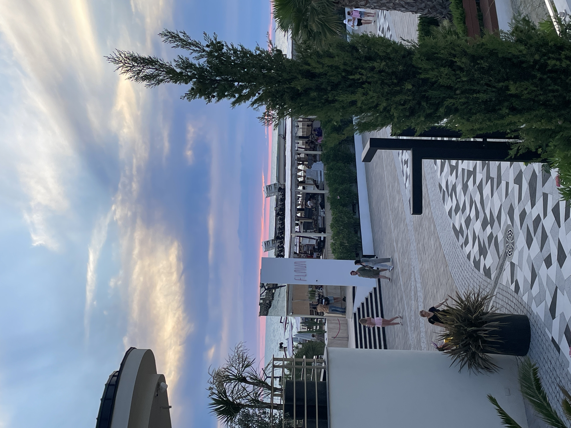 This image shows a sunset scene at a coastal establishment, likely a restaurant or beach club, in Sochi, Russia. The sky displays a gradient of colors, from light blue with white clouds to pink and purple hues near the horizon, indicating it is twilight. The sea is visible in the background, with a thin line of pink separating it from the sky.
In the foreground, a modern building with a white, curved facade is partially visible on the left. A dark, large planter with spiky green foliage stands in front of it. The ground is paved with a geometric pattern of grey, white, and black tiles.
Several people are scattered across the scene. On the paved area, a woman in a light pink outfit walks away from the camera, while a man and a woman in neutral-colored clothing walk towards the camera. Near the planter, a woman in a black top and a person wearing a pink outfit with bunny ears are present. Further back, near the building with the name "FLAVA" on a white pillar, a group of people are standing around a bar or counter area.
The establishment in the background features open-air seating under a white canopy, with tables and chairs visible. Palm trees and other green foliage are interspersed throughout the scene. A tall, slender evergreen tree stands prominently in the right half of the frame, partially obscuring the view of the establishment and the sky. The overall impression is a lively evening atmosphere at a seaside resort.
