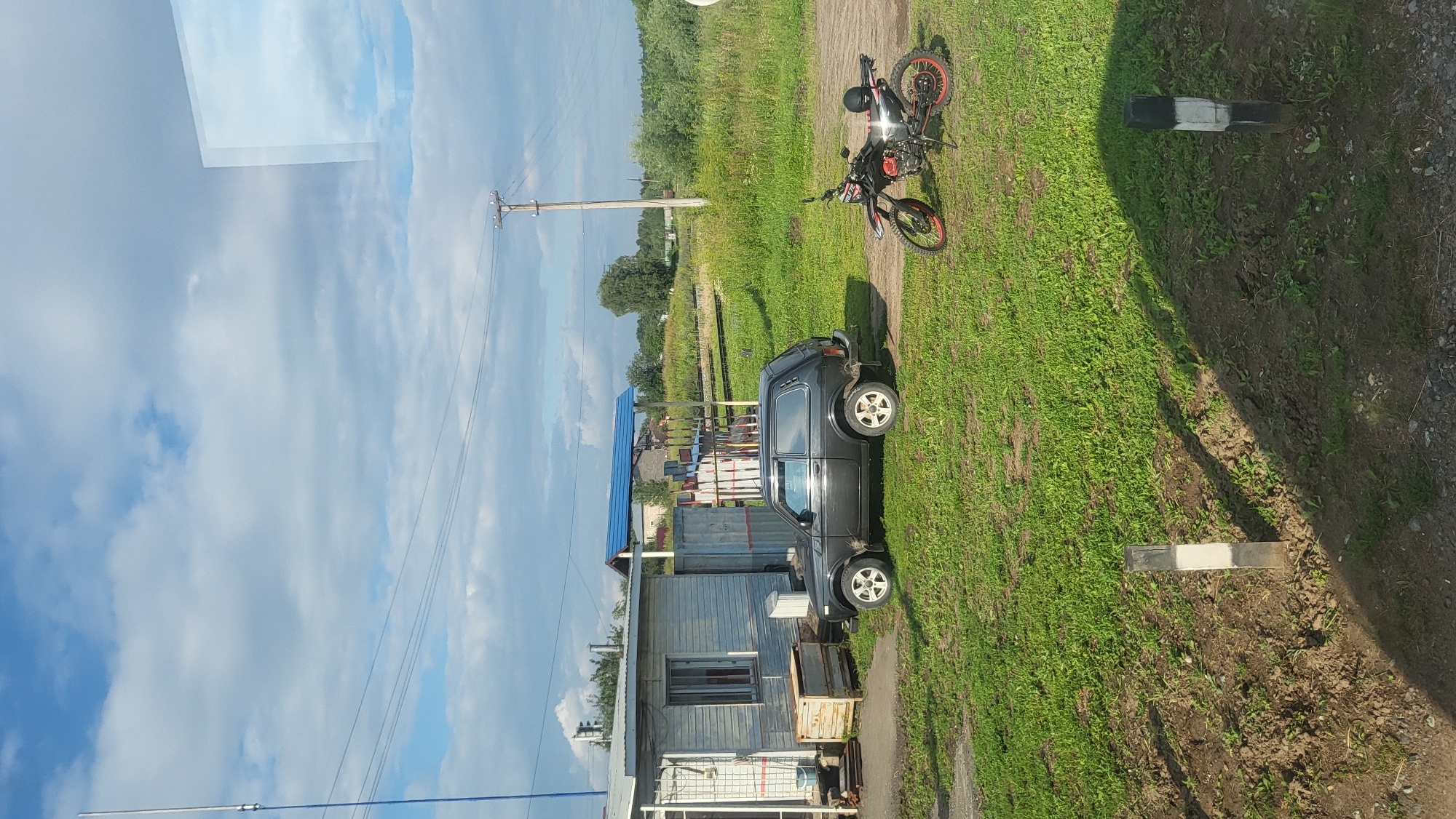 This is a ground-level, eye-level, vertical, full shot of a rural scene in Kvapsa, Russia. The image shows a dark-colored, three-door SUV parked on a dirt and grass lot. To the right of the SUV, a black dirt bike with red accents is parked on the same lot. A small, utilitarian structure, possibly a shed or outbuilding, is located to the left of the SUV. A blue awning extends from this structure. The background consists of green grass, trees, and a cloudy sky. In the foreground, two striped posts, one black and white, the other black and white, are visible in the dirt. The scene appears to be an outdoor, undeveloped area.