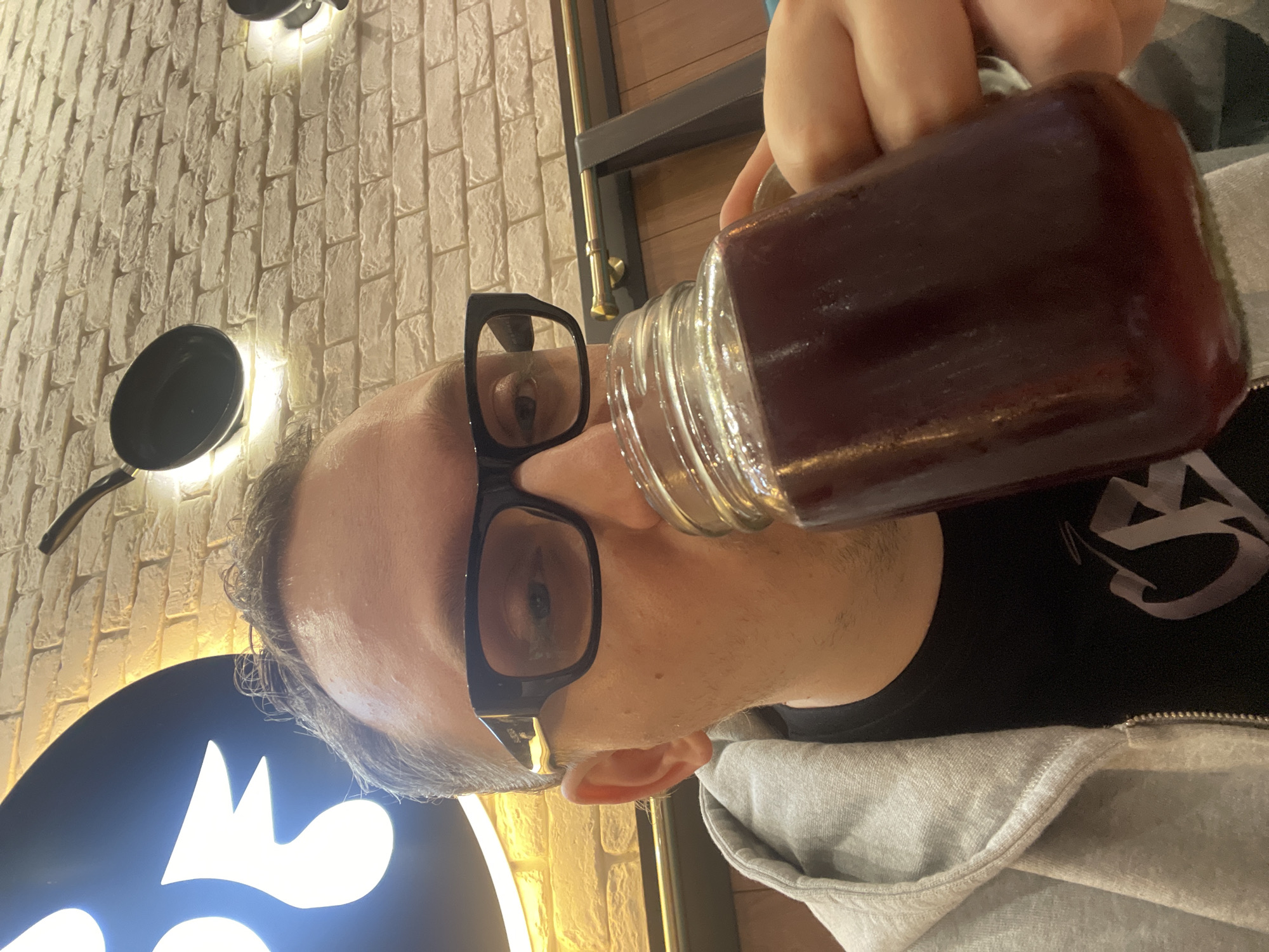 A person is drinking from a glass jar of dark liquid. The person is wearing glasses and a gray hooded sweatshirt. The background consists of a brick wall with some lights and a black sign with a white illuminated design. The image appears to be taken indoors during the day. The person's expression is neutral, and the overall mood is casual. There is no visible text in the image.